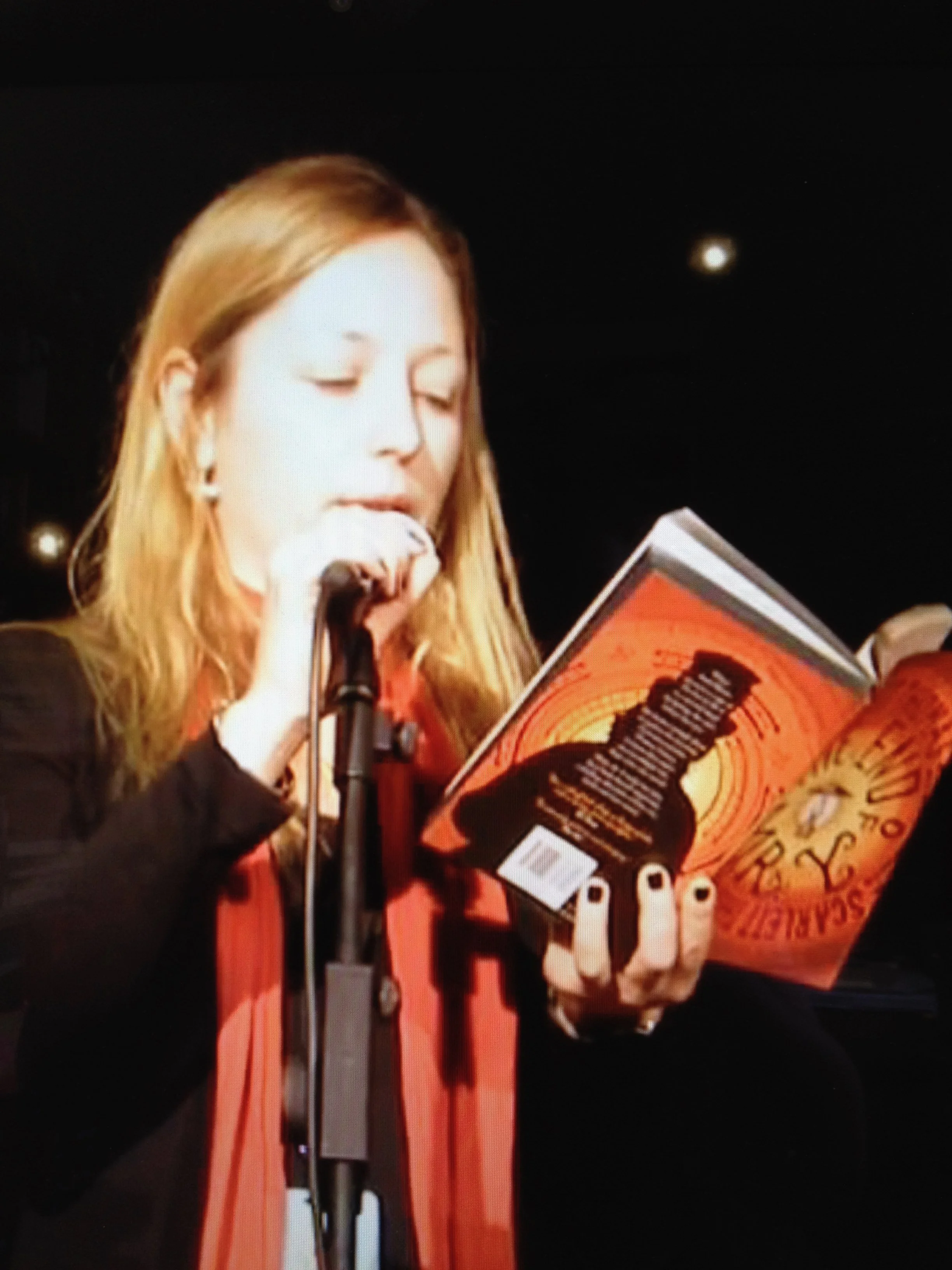 A woman with light brown hair reading from a book in front of a microphone.