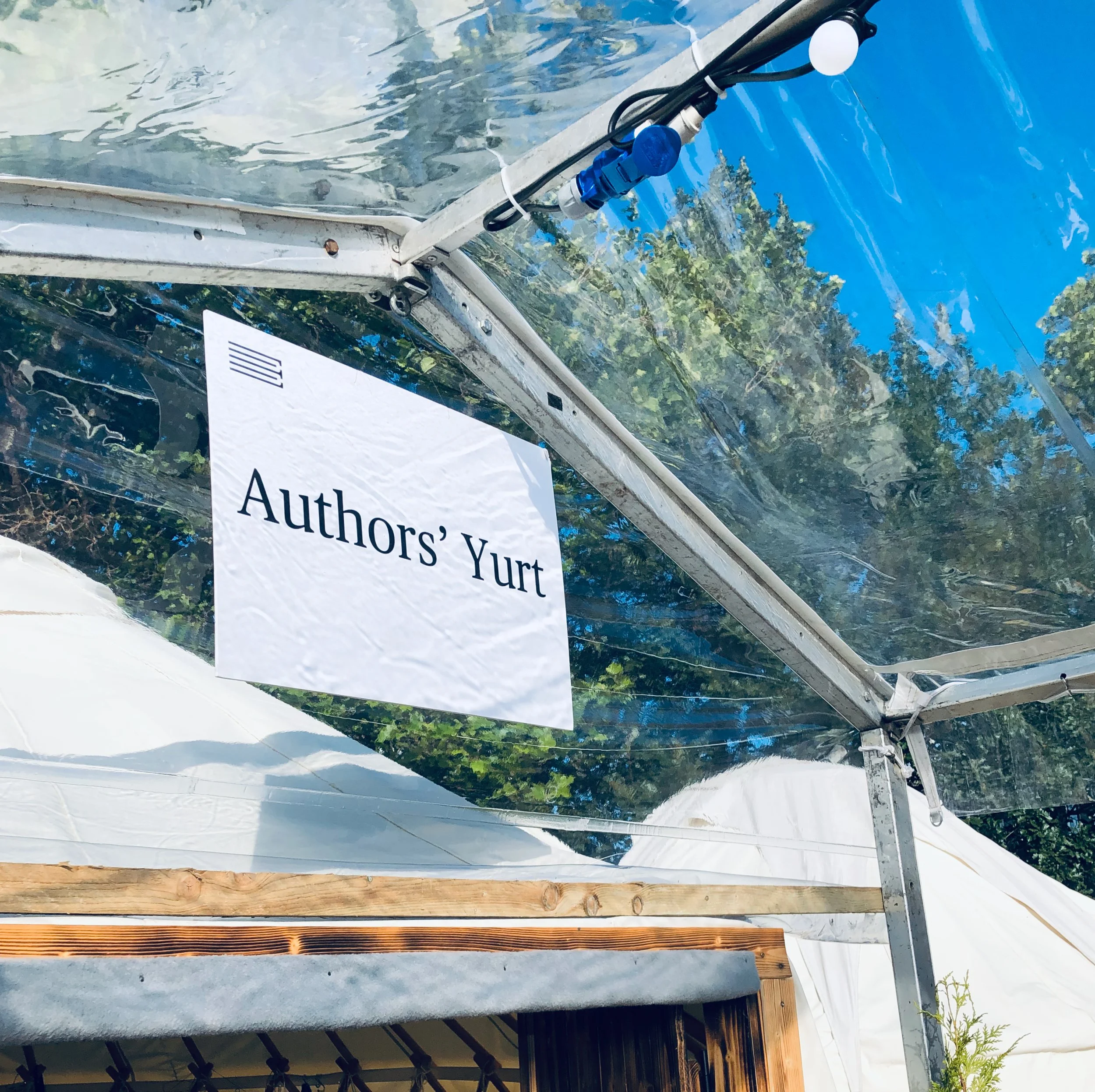 A sign inside a greenhouse or tent that reads 'Authors' Yurt' with transparent plastic walls and a wooden structure, with trees and blue sky visible outside.