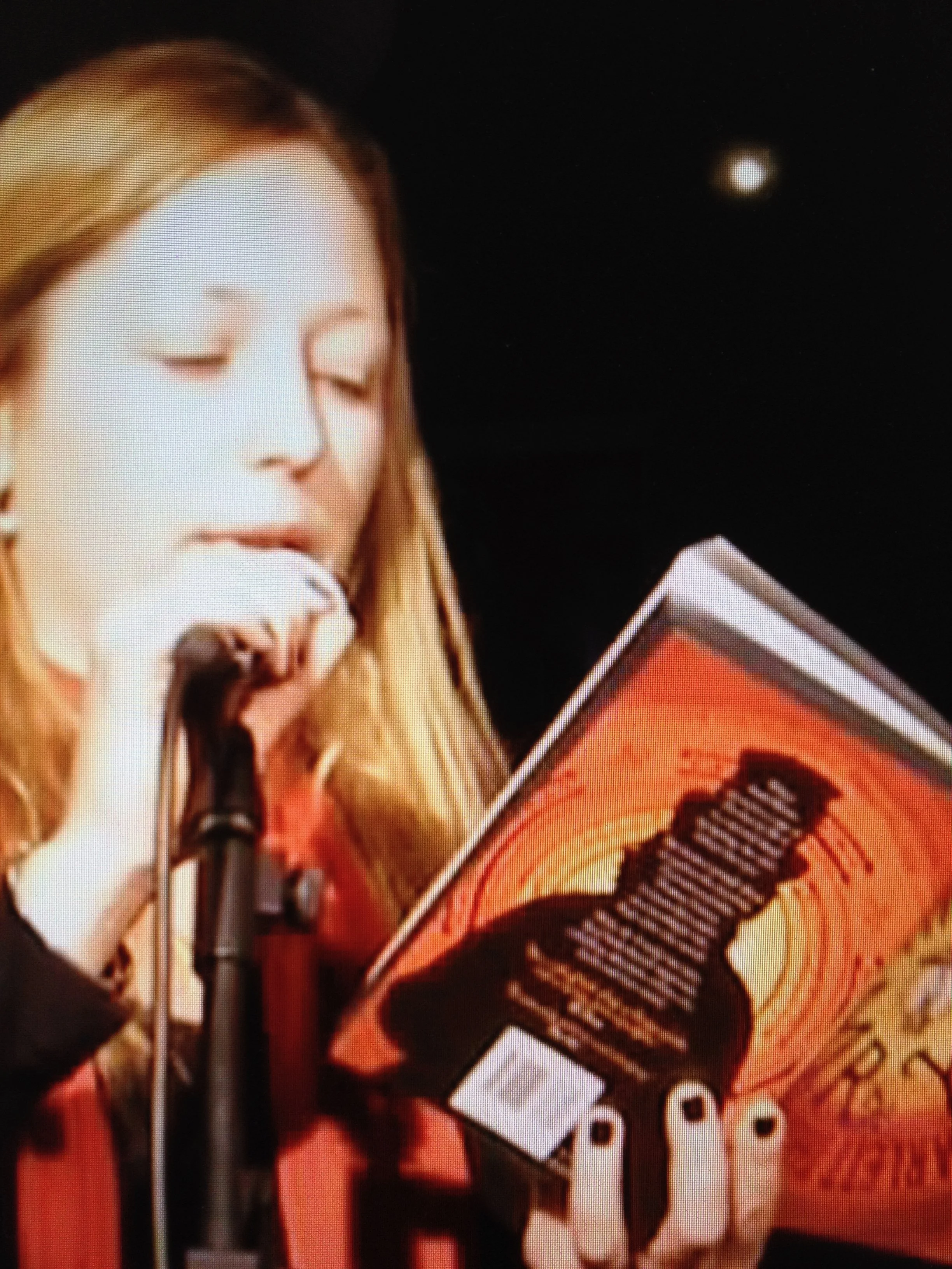A woman with long, light brown hair reading a book titled 'The Handmaid's Tale' while standing at a microphone on a stage with a dark background and a small bright light or star.