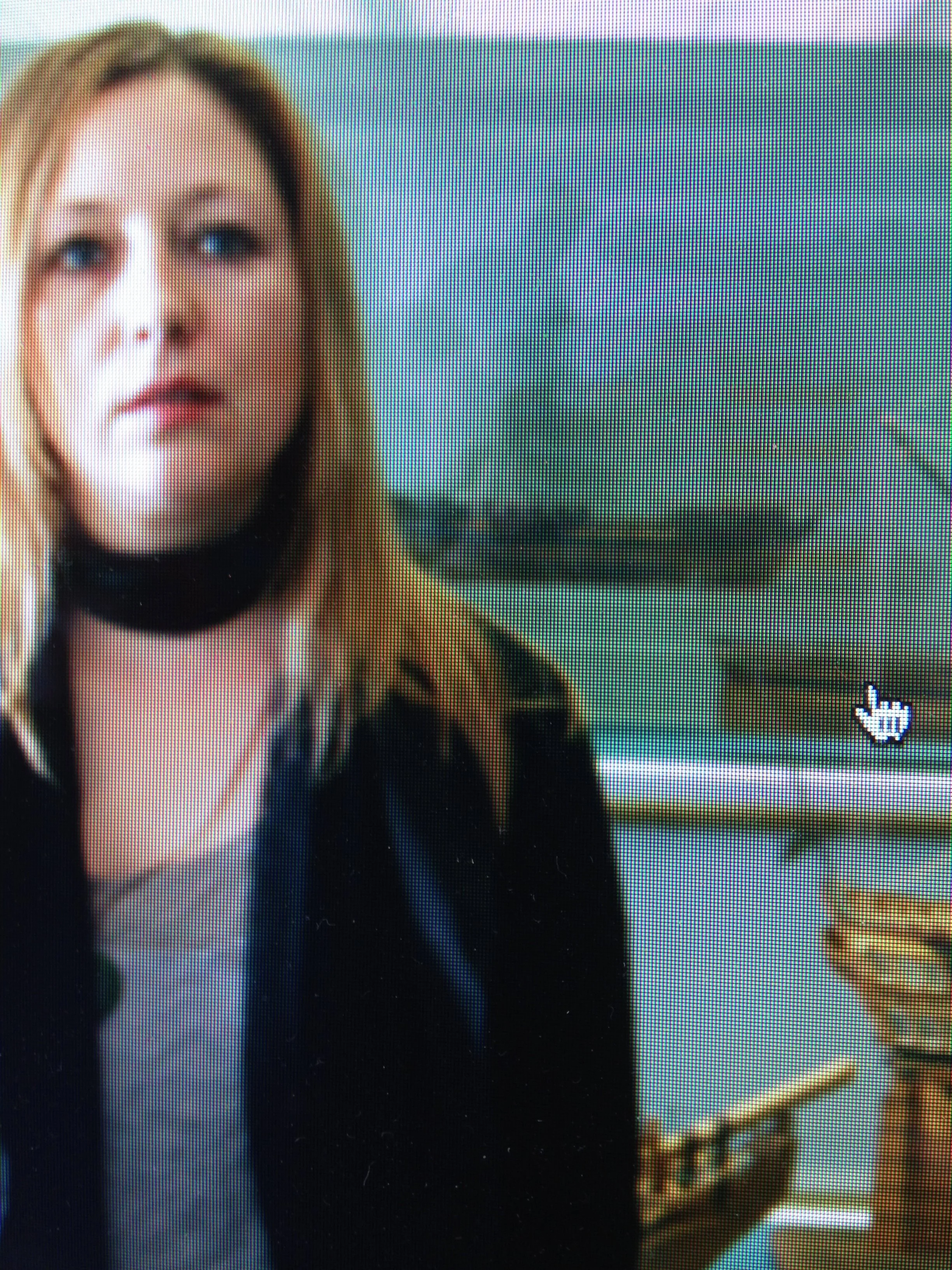 A woman with shoulder-length blonde hair wears a black choker and black jacket, standing outdoors with a green landscape in the background.