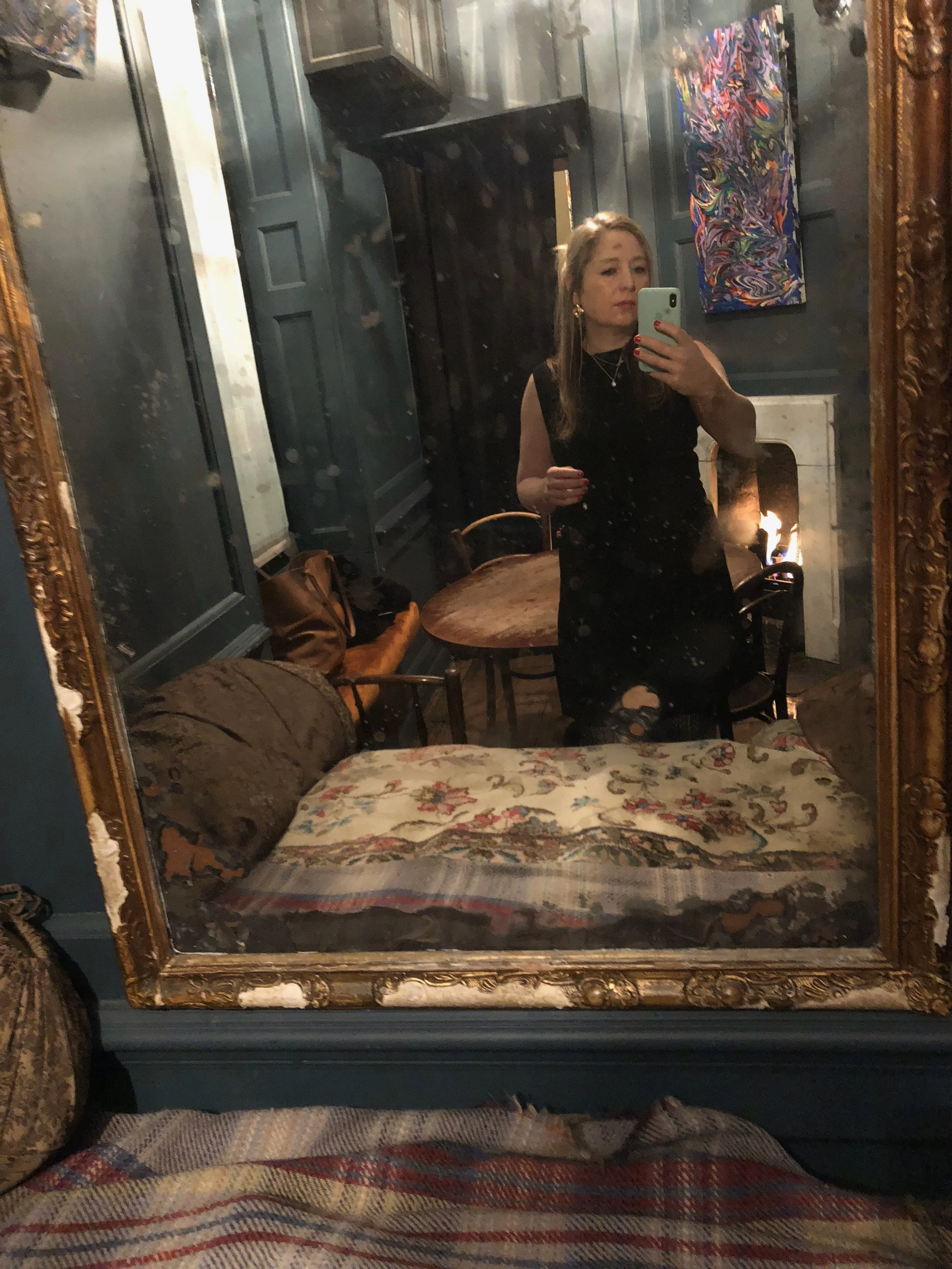 A woman taking a selfie in a large, ornate mirror in a cozy, vintage-style room with a fireplace, colorful painting, and furniture.