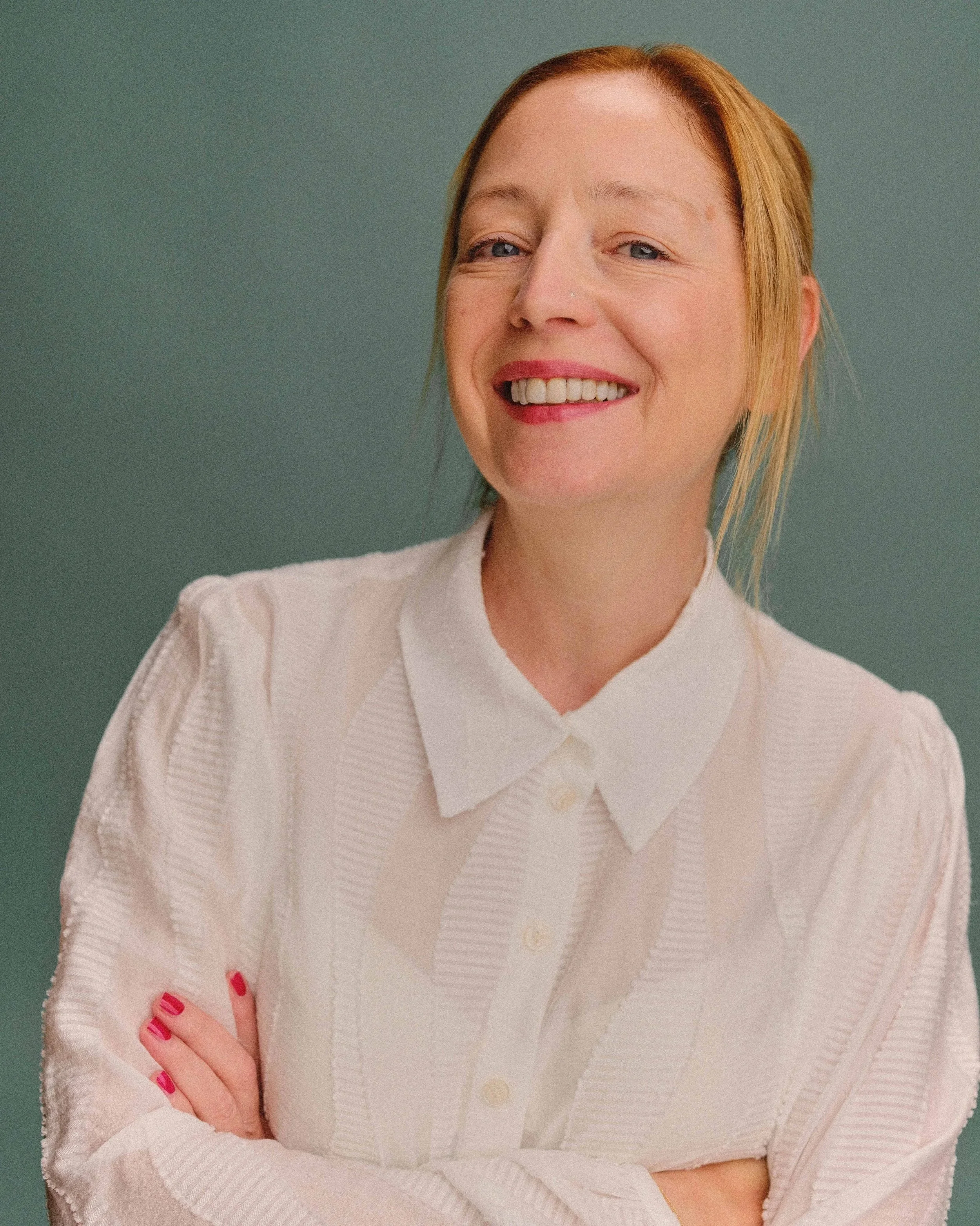 Storyteller Scarlett Thomas smiling, wearing a white button-up shirt, with her arms crossed, standing against a muted green background.
