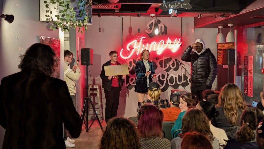 People performing on a small stage at a venue, with one woman singing into a microphone, surrounded by an audience, and a neon sign that says 'I'm happy to see you!'