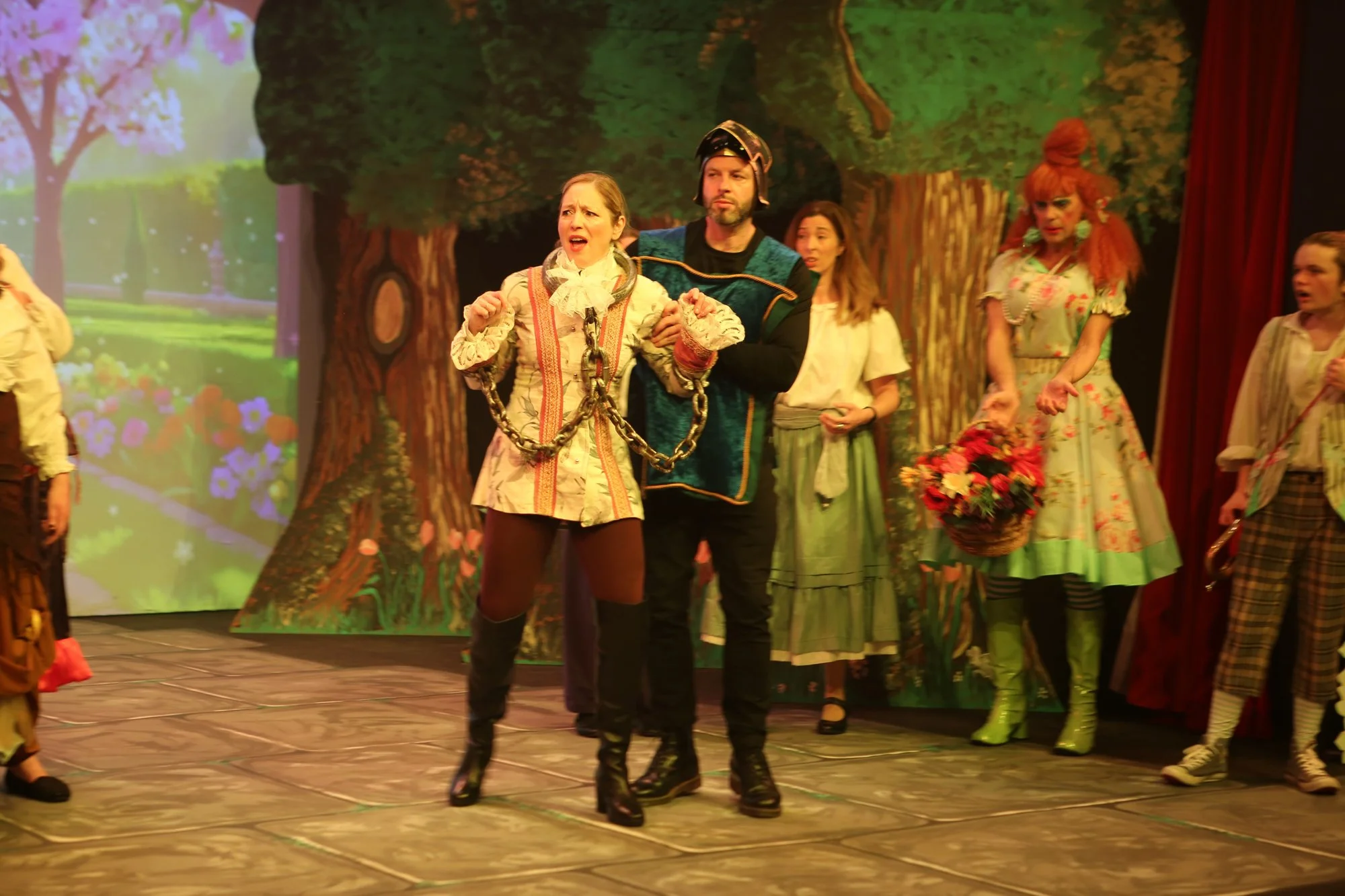 Actors performing in a theatrical play set in a forest, with characters in colorful costumes, including a woman with chains and a man dressed as a soldier, on stage with a backdrop of trees and flowers.