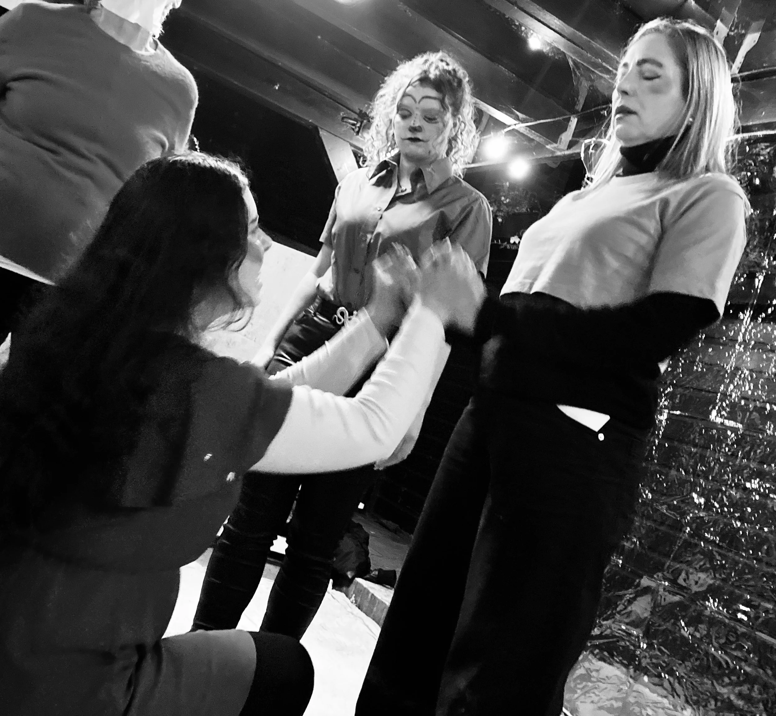 Four women engaging in a dramatic conversation indoors, with one woman kneeling and touching the hand of a standing woman, while a third woman with curly hair appears concerned and a fourth woman stands nearby.