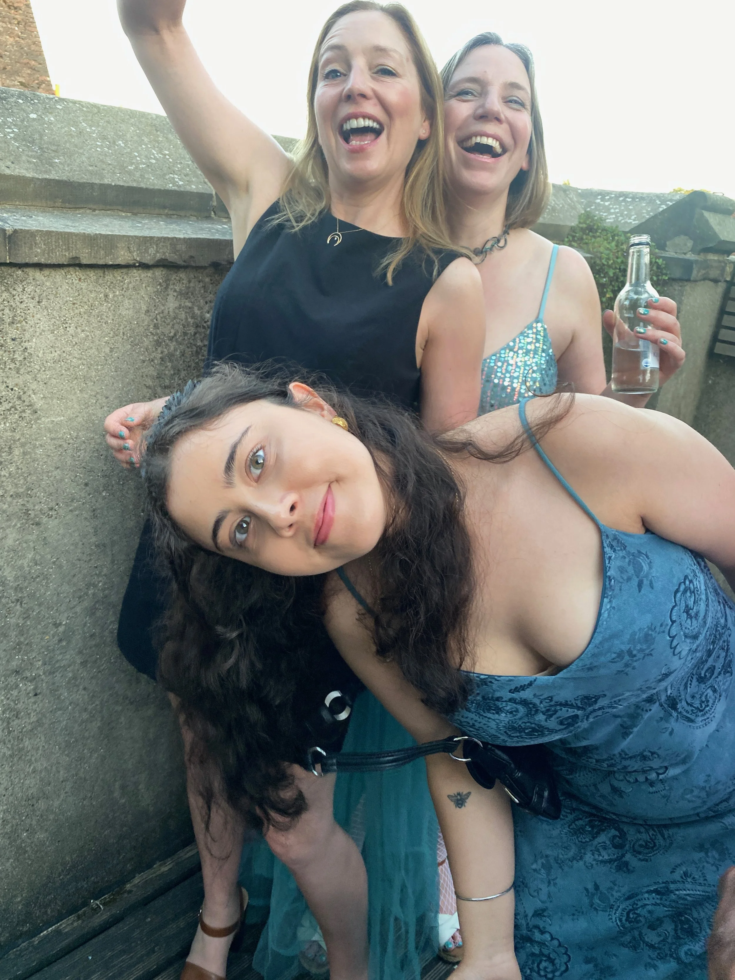 Four women posing together outside. One woman is lying down in front, wearing a blue patterned dress. Behind her, three women are standing, smiling, and making expressions of joy, with shared laughter. One woman has long red hair, another has short b