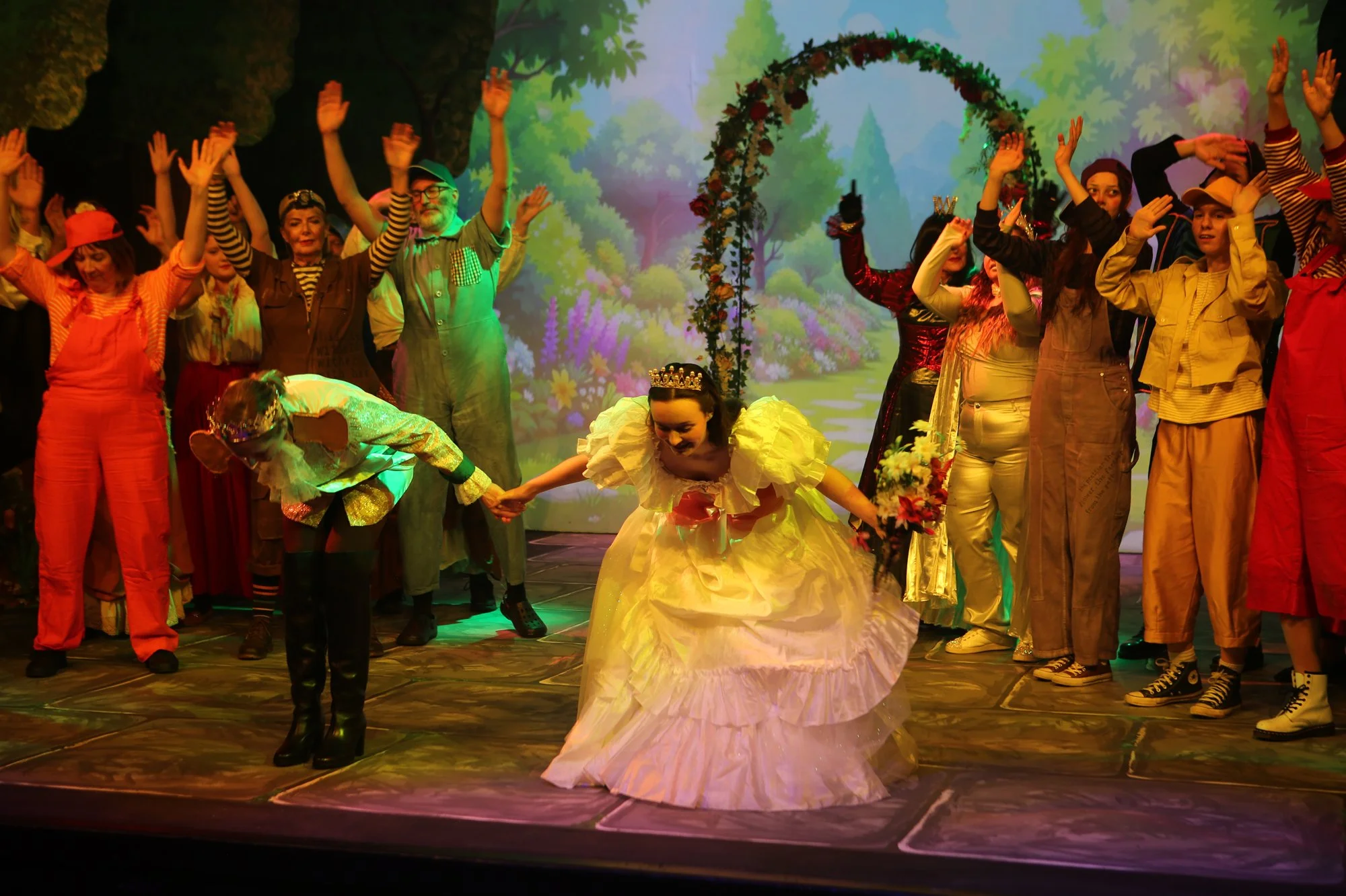 The image depicts a theatrical stage scene with performers dressed in colorful costumes. The central figure is a woman dressed as a princess holding a bouquet with other actors around her, all with their hands raised. The background features a painte