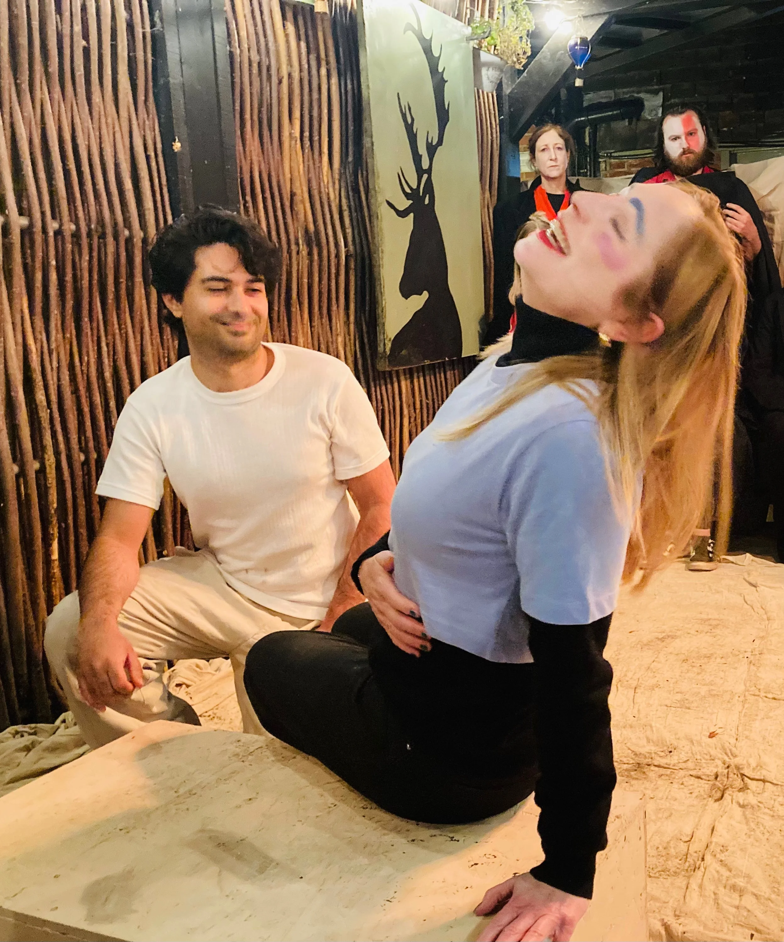 A woman with blonde hair, wearing makeup, leaning back with her eyes closed, and a man with dark hair and a white T-shirt smiling and sitting nearby. Two other people are standing in the background, one with glasses and a beard, and a painting of a d