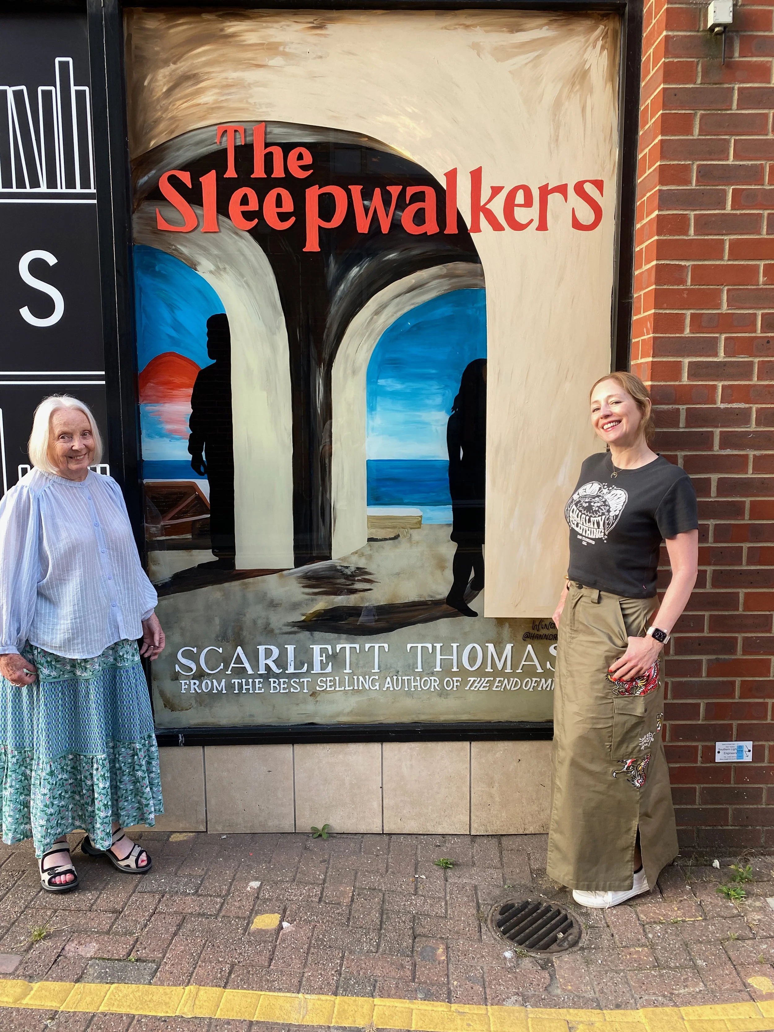 Two women standing in front of a painted theater poster for 'The Sleepwalkers' by Scarlett Thomas. The woman on the left has white hair and is wearing a light blue striped blouse, floral skirt, and sandals. The woman on the right has light brown hair