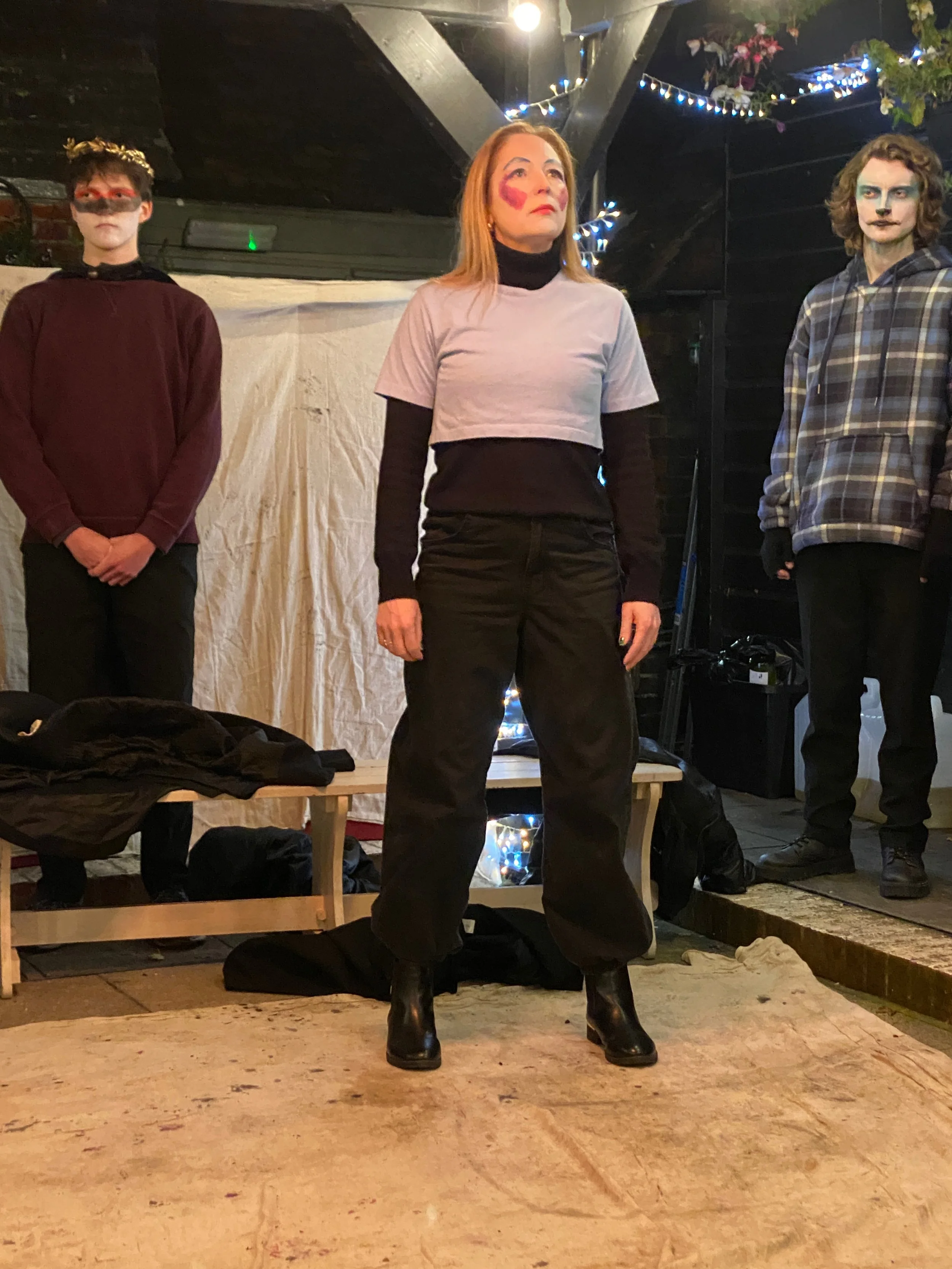Three people dressed in casual clothing with face paint, standing on a stage with festive string lights and flowers above. The woman in the center has heart-shaped face paint and stands with her hands at her sides. The two young men on either side ha