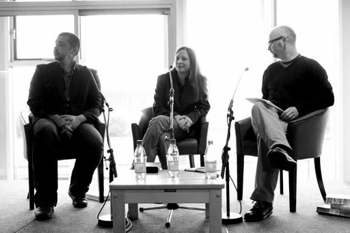Three people sitting in chairs on a panel discussion or interview, with microphones in front of them, in a well-lit room with large windows.
