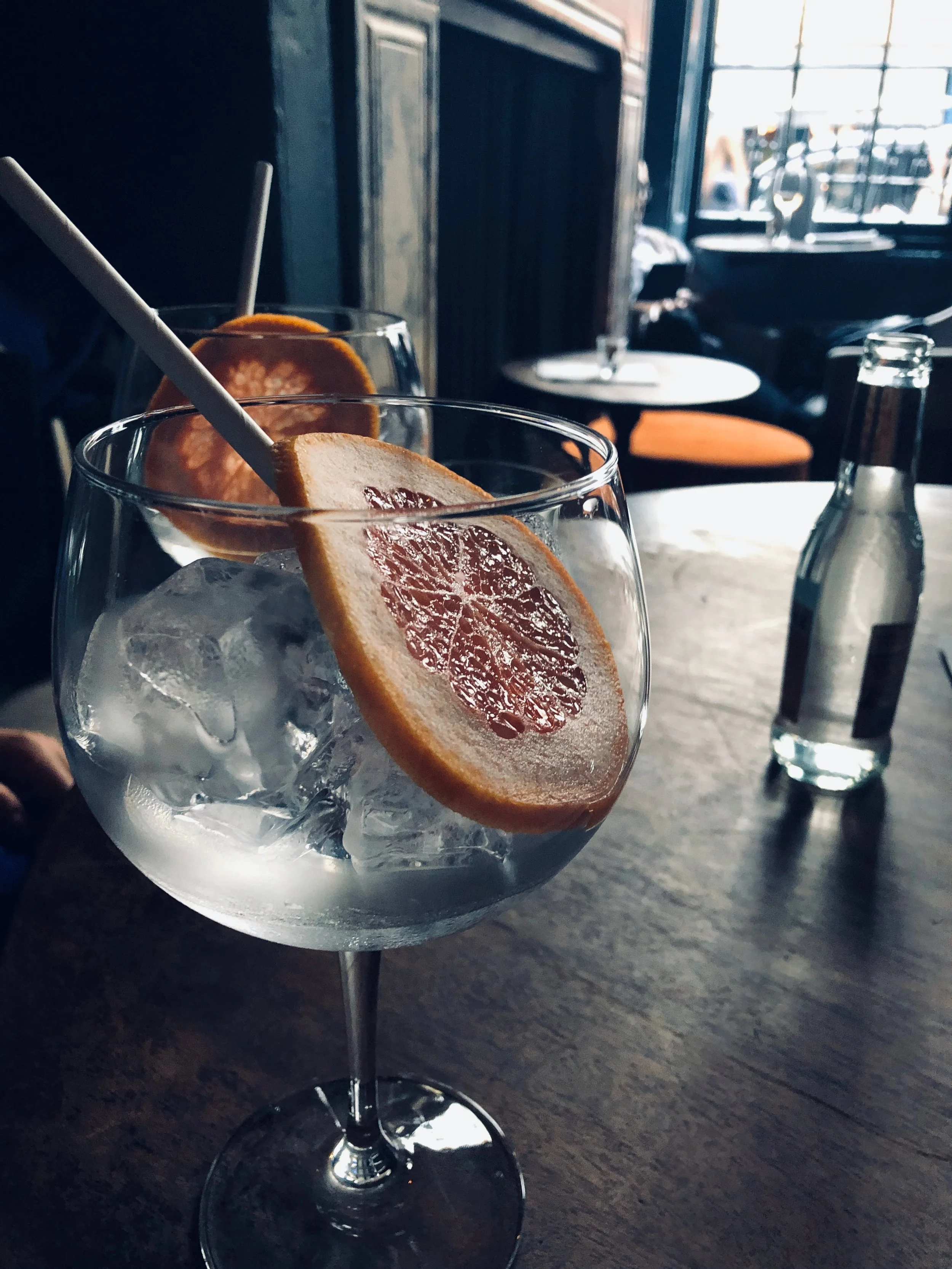 A cocktail glass filled with ice and garnished with a dried orange slice, on a wooden table in a dimly lit cafe or bar.