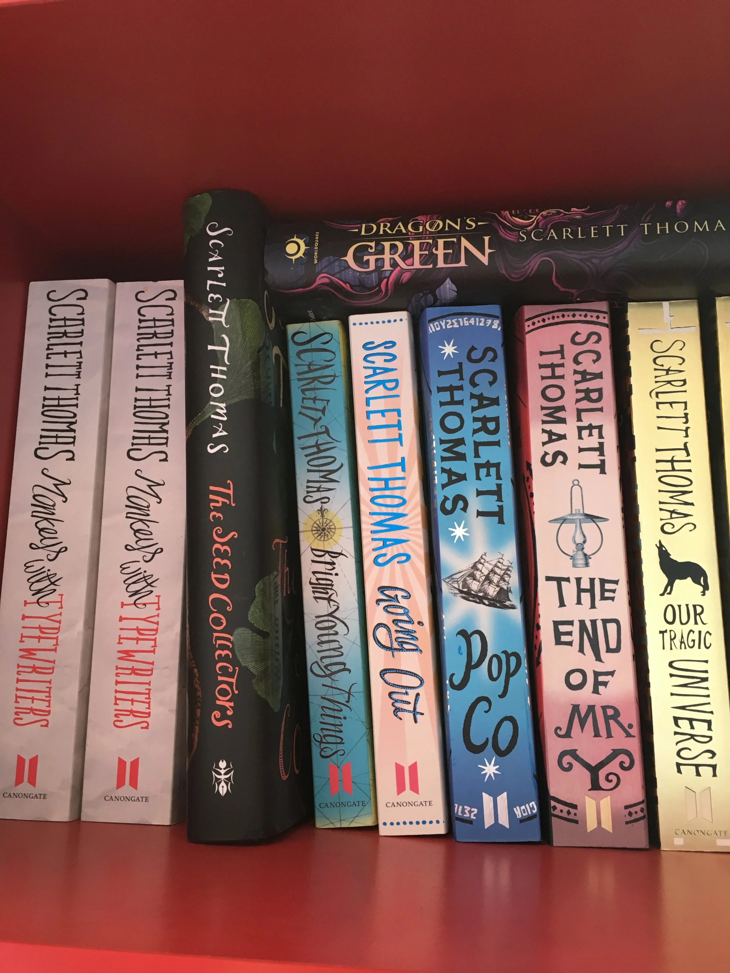 A collection of romance novels on a shelf, including titles like 'Scarlett Thomas', 'Dragon's Green', 'The End of Mr. Y', and 'Our Tragic Universe'.