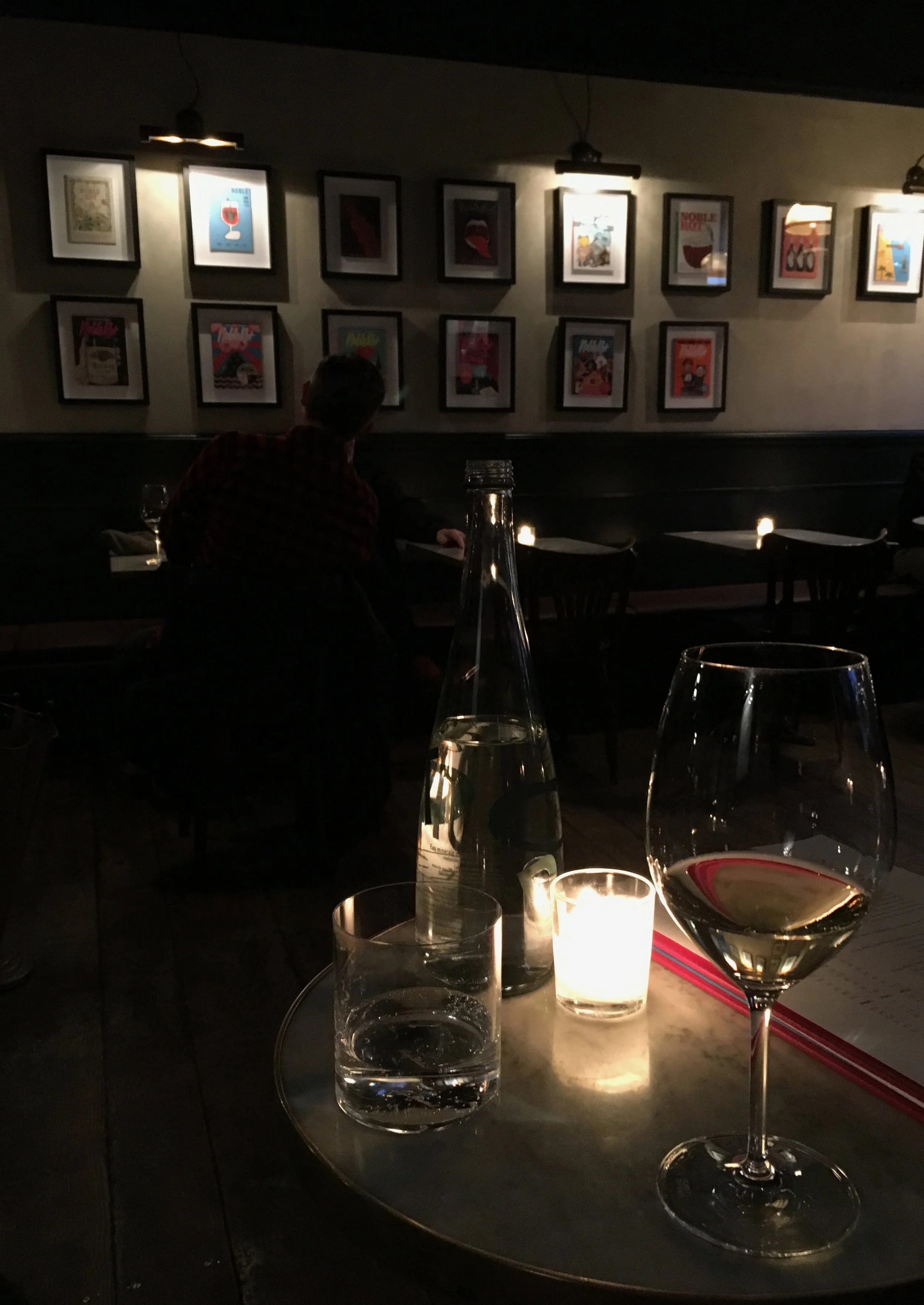 A dimly lit restaurant or bar with framed artwork on the wall, a man sitting at a table, glass of white wine, bottle of water, and lit candle on a round table in the foreground.