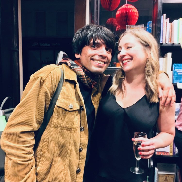 A man and woman smiling and hugging at an indoor party, woman holding a glass of champagne, with red paper lanterns and books in the background.