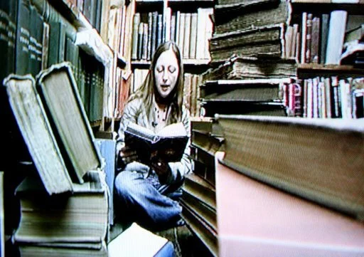A woman sitting in a small library surrounded by shelves filled with books, reading a book.
