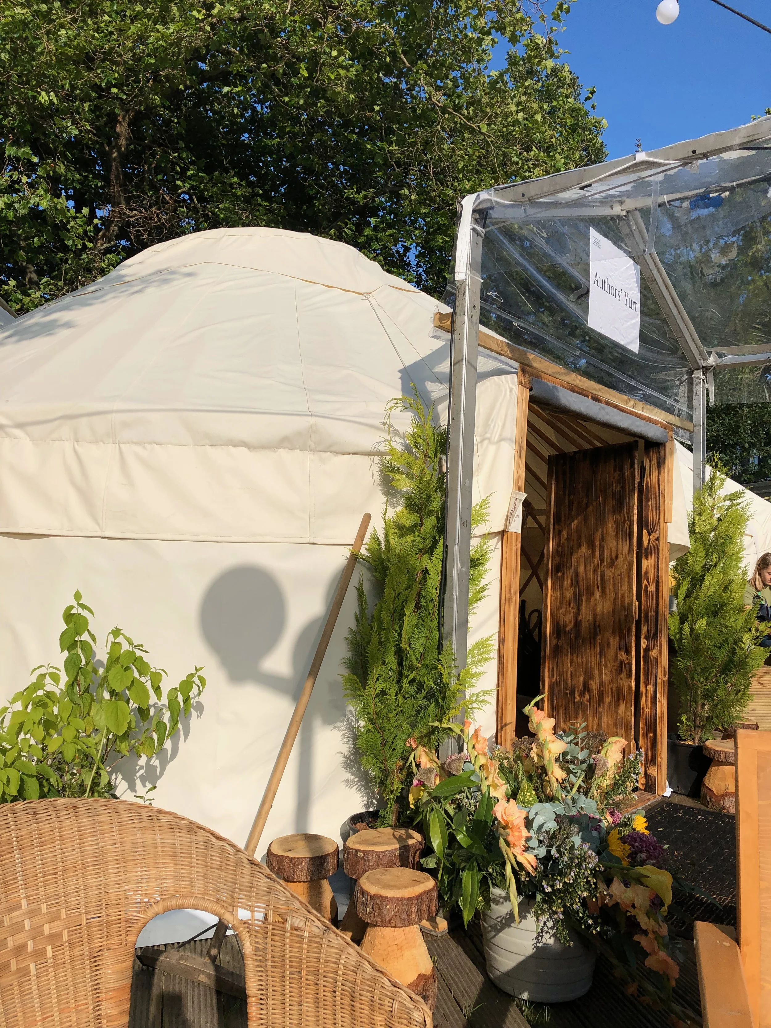 A white yurt with a wooden door, surrounded by potted plants and flowers, under a blue sky with trees in the background. A sign on the clear plastic panel says "Author's Yurt."