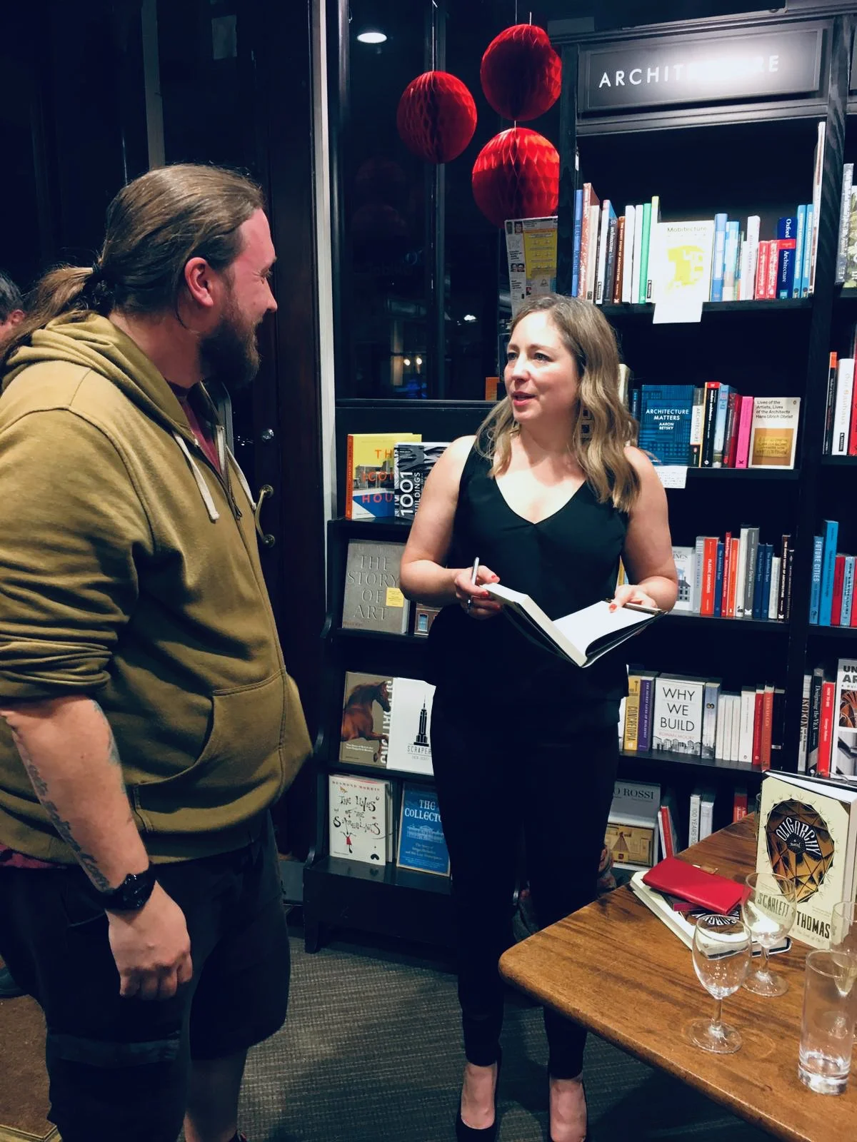 A woman with wavy blonde hair holding a book and talking to a man with long hair and a beard, wearing a brown hoodie, inside a bookstore or library with shelves of books and decorative red lanterns overhead.