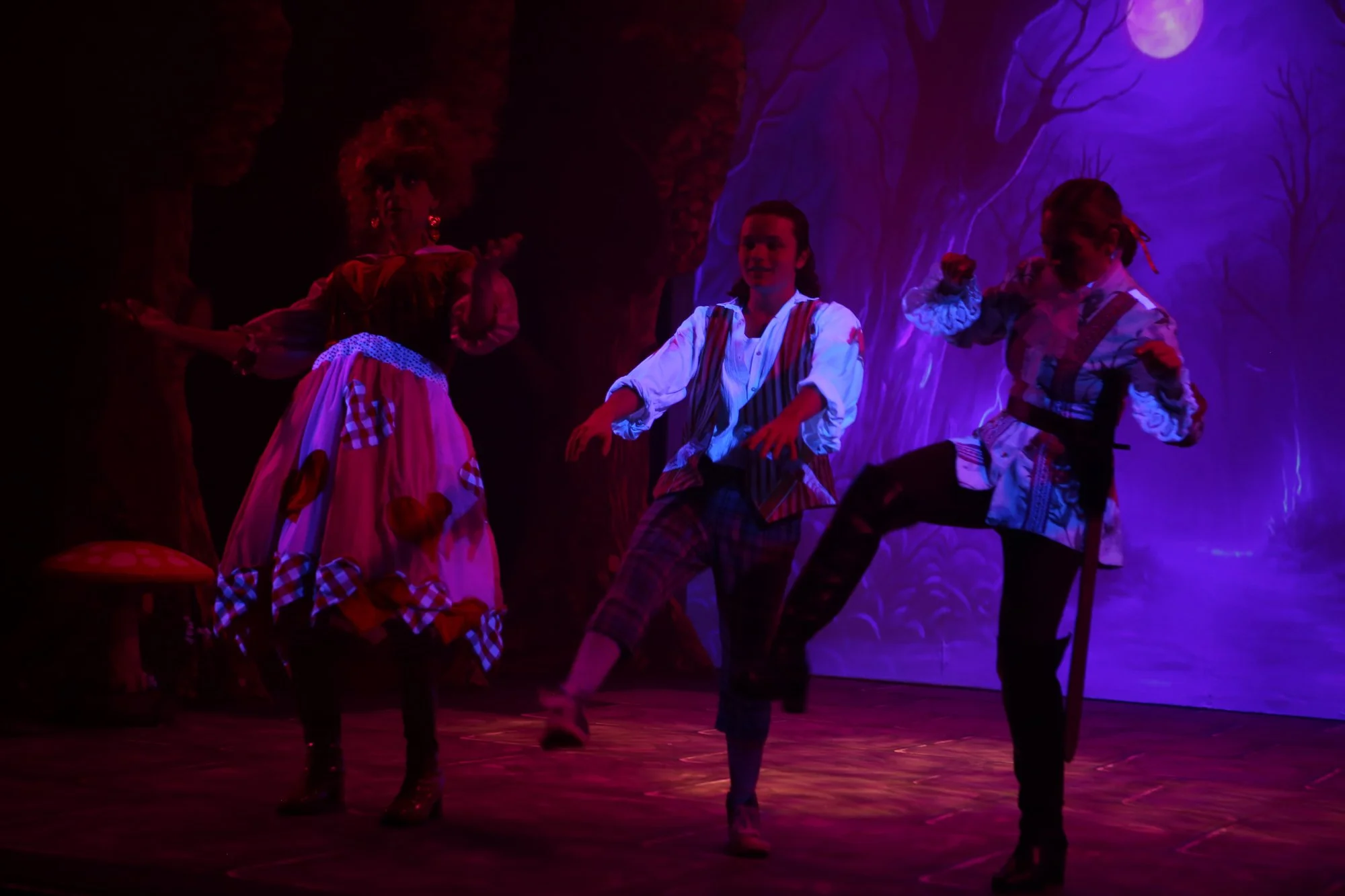 Three performers in costumes dancing on a stage with a colorful forest backdrop, under purple stage lighting.