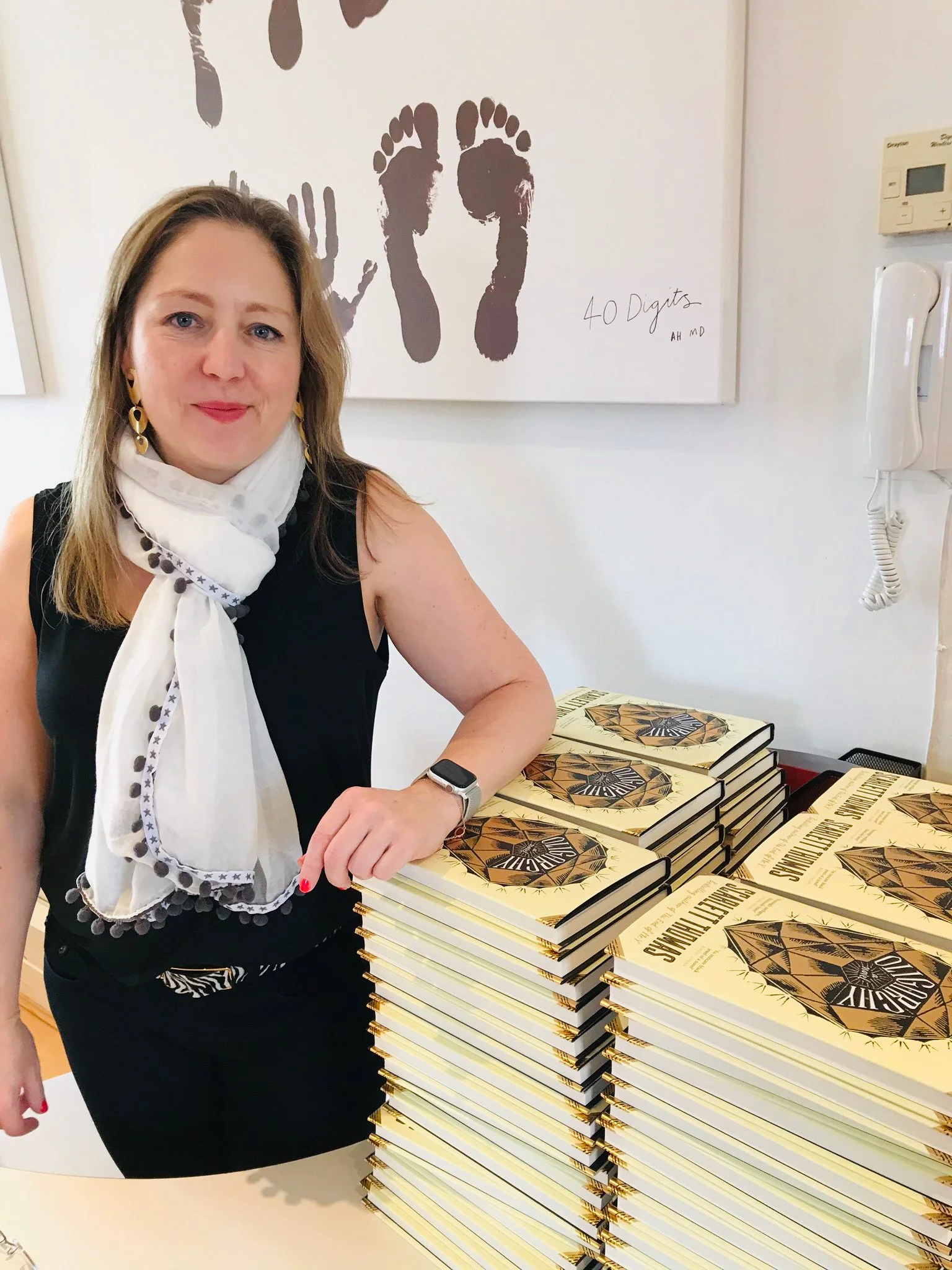 A woman standing next to a large stack of books titled "Slavery in the United States" in a room with a wall art of two dark footprints and the text "40 Digits".