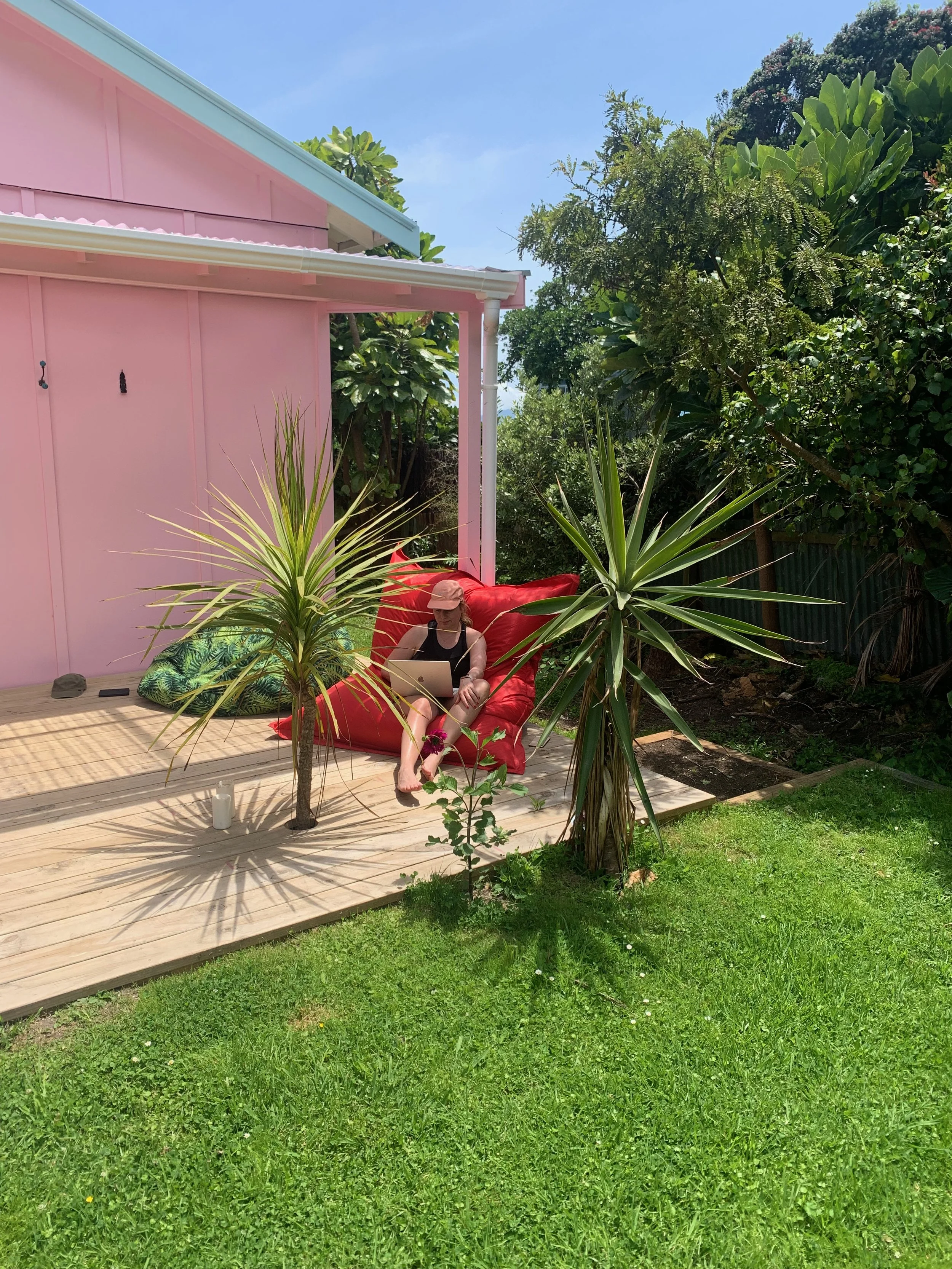 A person sitting on a red outdoor chair with a laptop in a backyard, surrounded by green plants, a wooden deck, and a pink house with trees in the background.