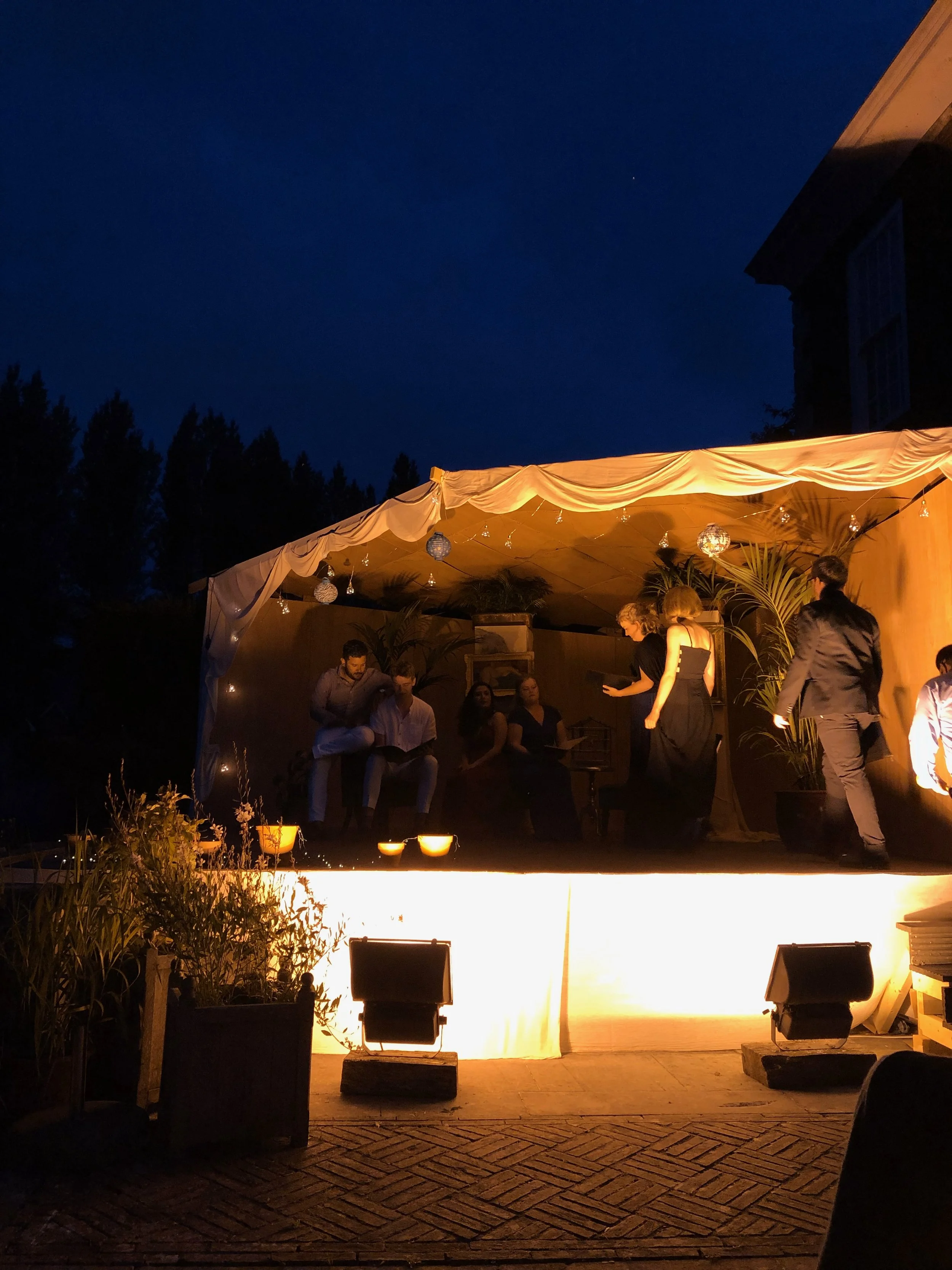 People socializing and dancing on an outdoor stage with warm lighting at nighttime.