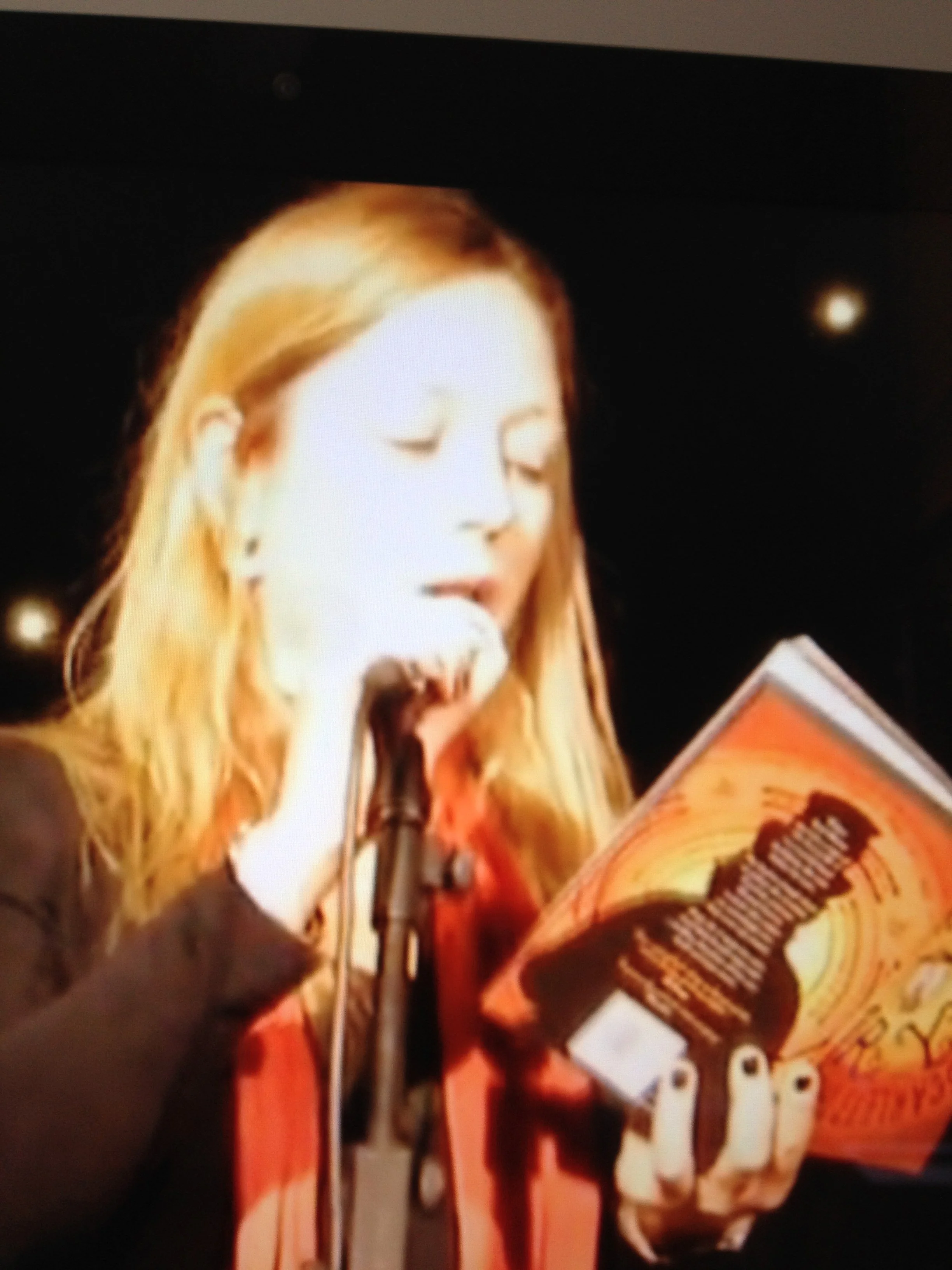 A woman with blonde hair reading from a book into a microphone on a dark background.