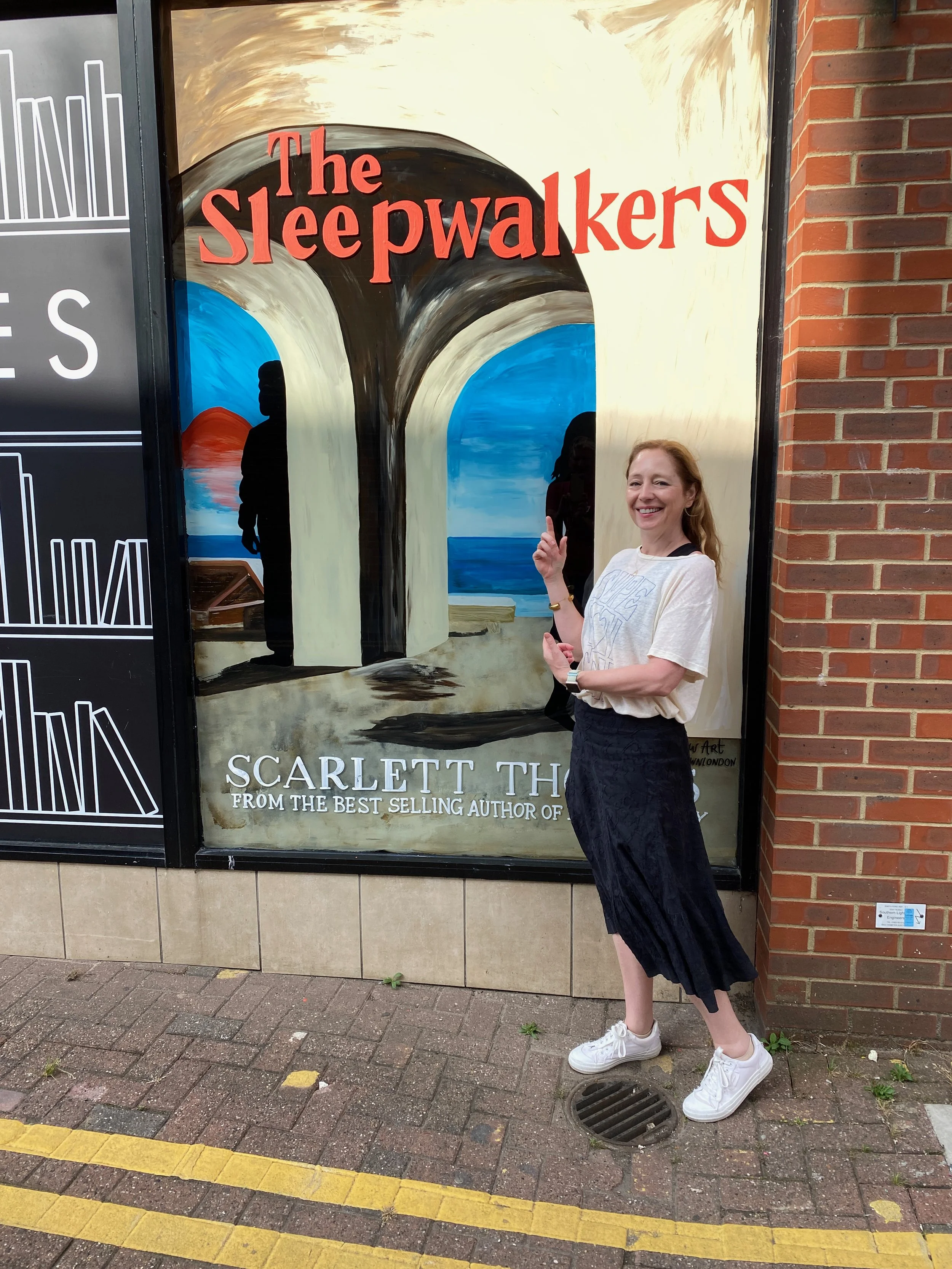Woman standing in front of a poster for the book "The Sleepwalkers" by Scarlett Th