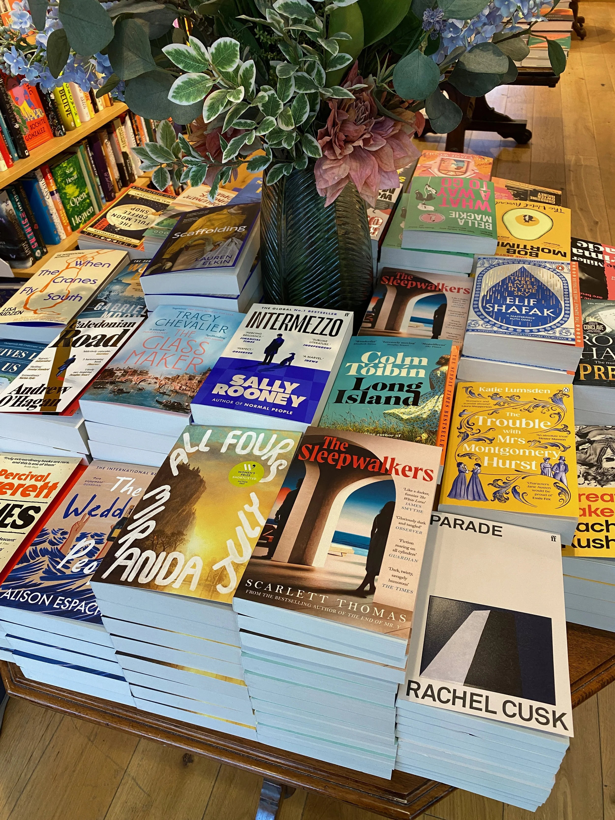 Stack of books on a table with a vase of flowers on top, surrounded by more books and bookshelves in the background.