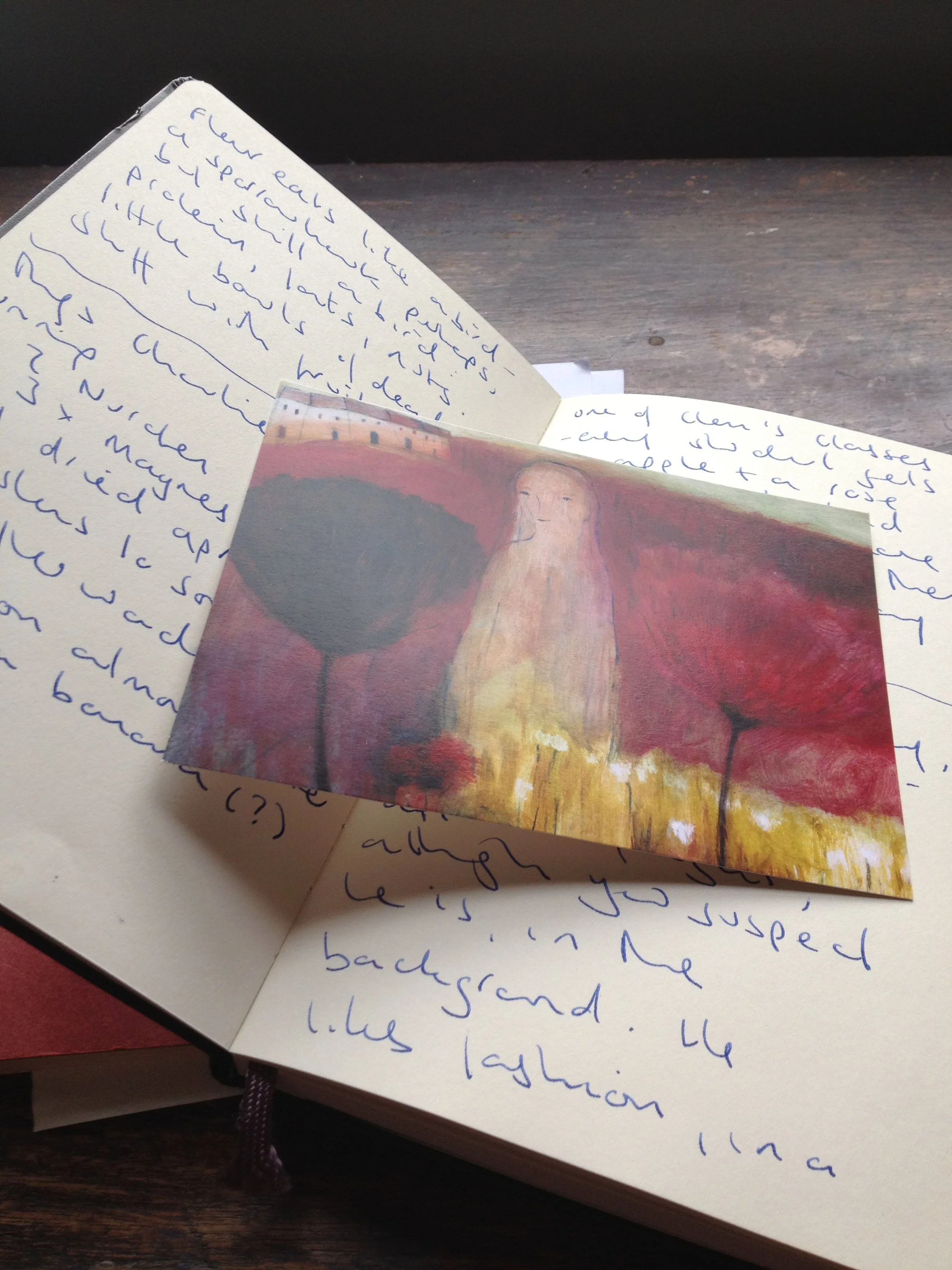 Open notebook with handwritten notes in blue ink, partially covering a small painting or reproduction of a painting featuring a woman with long hair and abstract red and black trees.