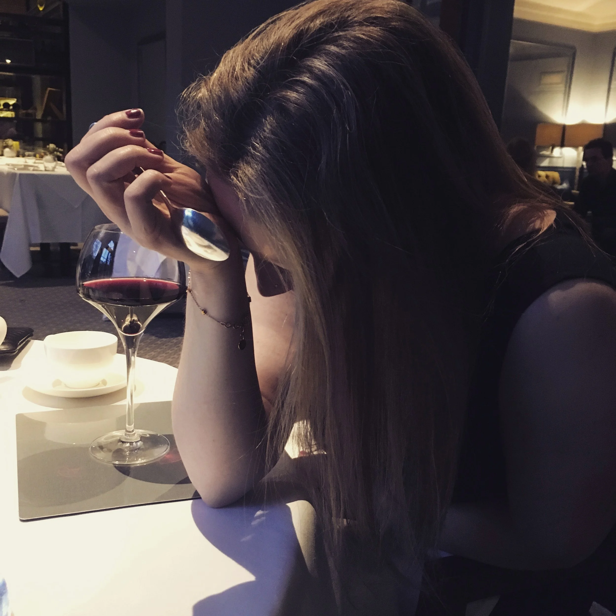 A woman sitting at a restaurant table with her head resting on her hand, holding a spoon, and a glass of red wine nearby.