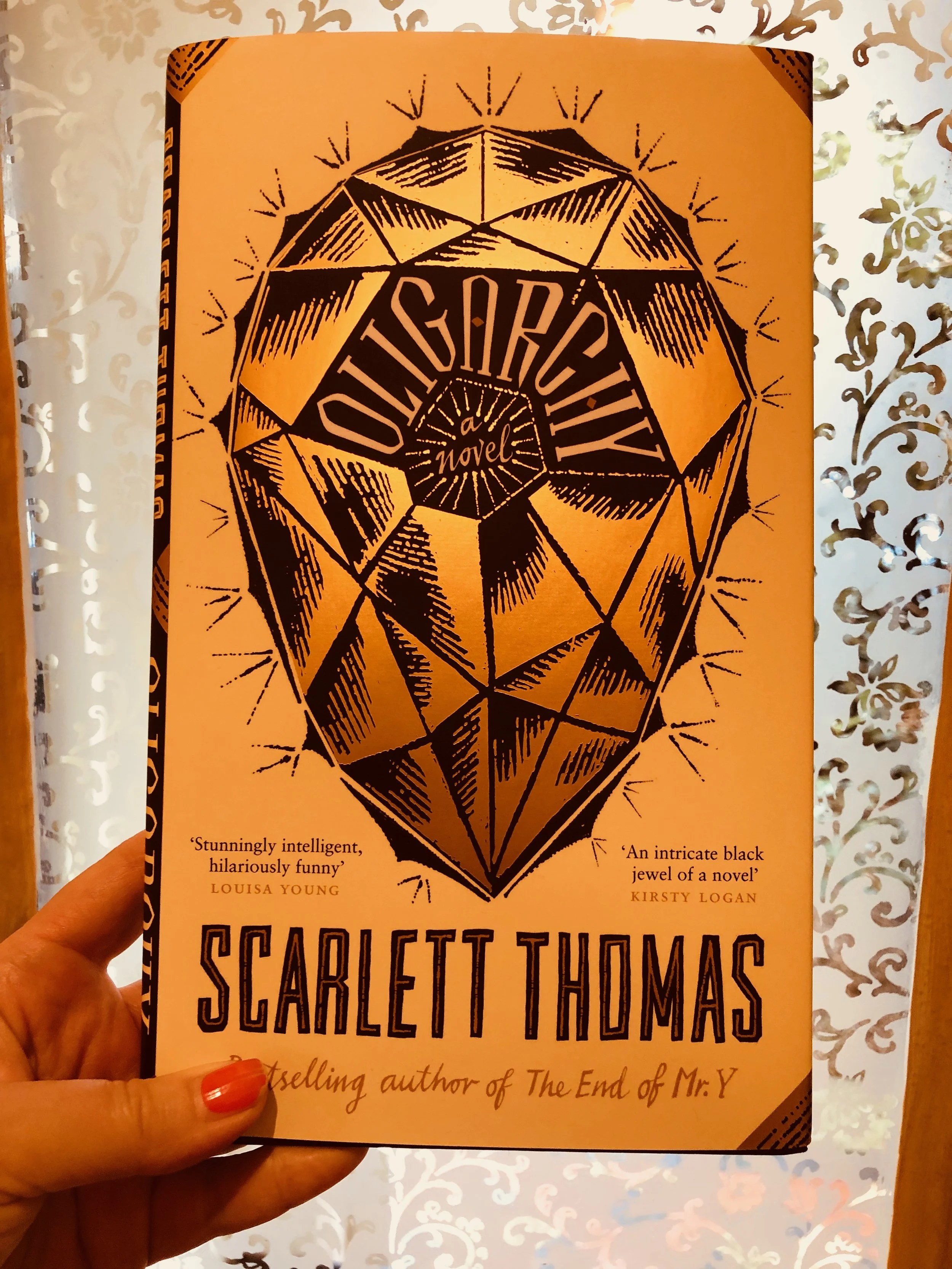 A person holding a paperback book titled 'The Seven Husbands of Evelyn Hugo' by Scarlett Thomas, with a black and orange geometric gemstone illustration on the cover. The background features a decorative white patterned curtain or wallpaper.