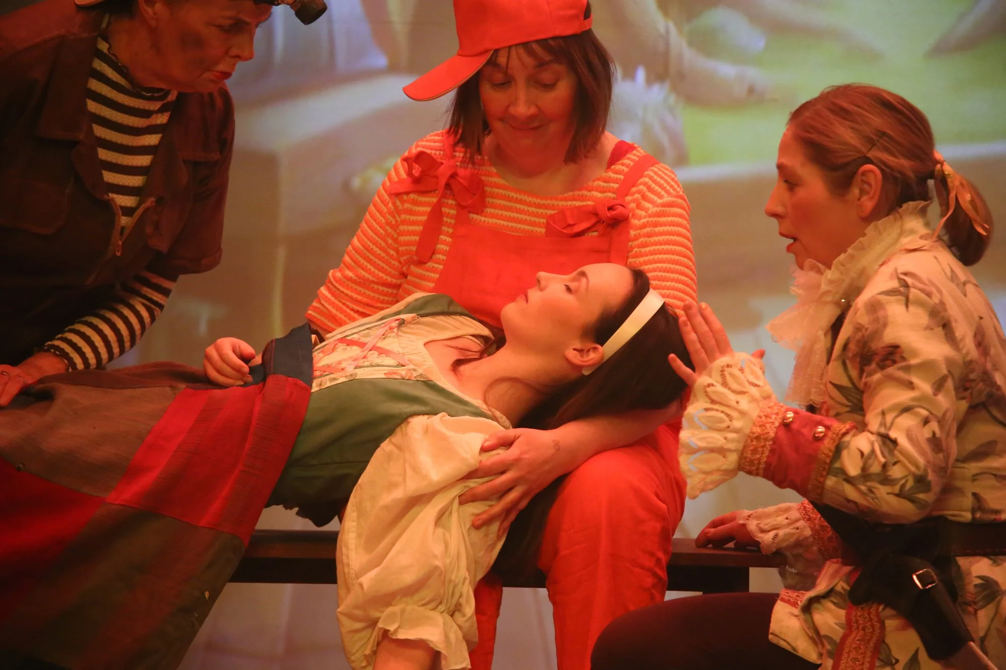 A woman lying on a table with her head in the lap of another woman, surrounded by three women all looking at her calmly.