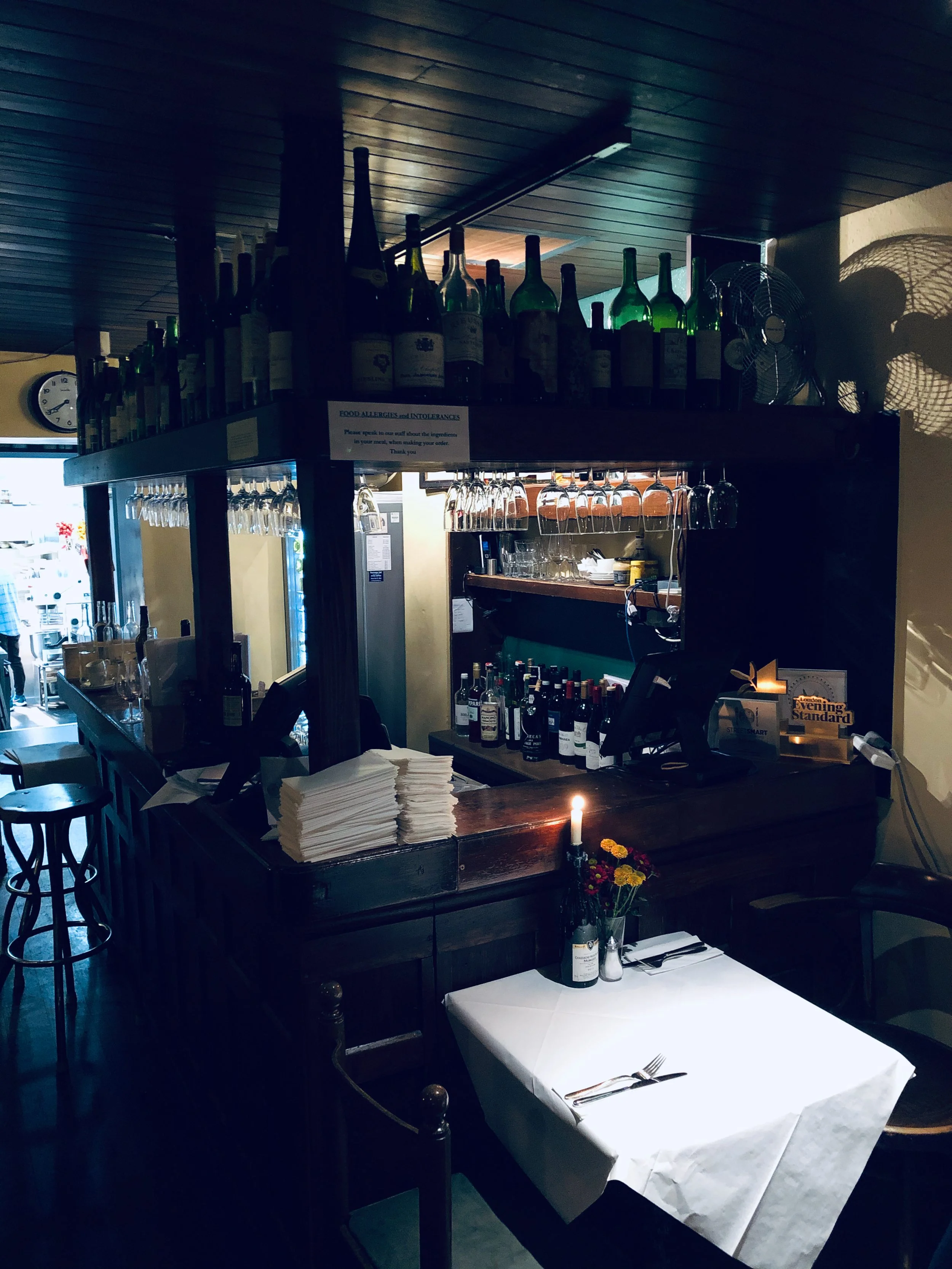 A cozy restaurant interior with a bar counter stocked with wine bottles and glasses, a small table set for dining with a white tablecloth, a candle, a flower vase, and place settings.