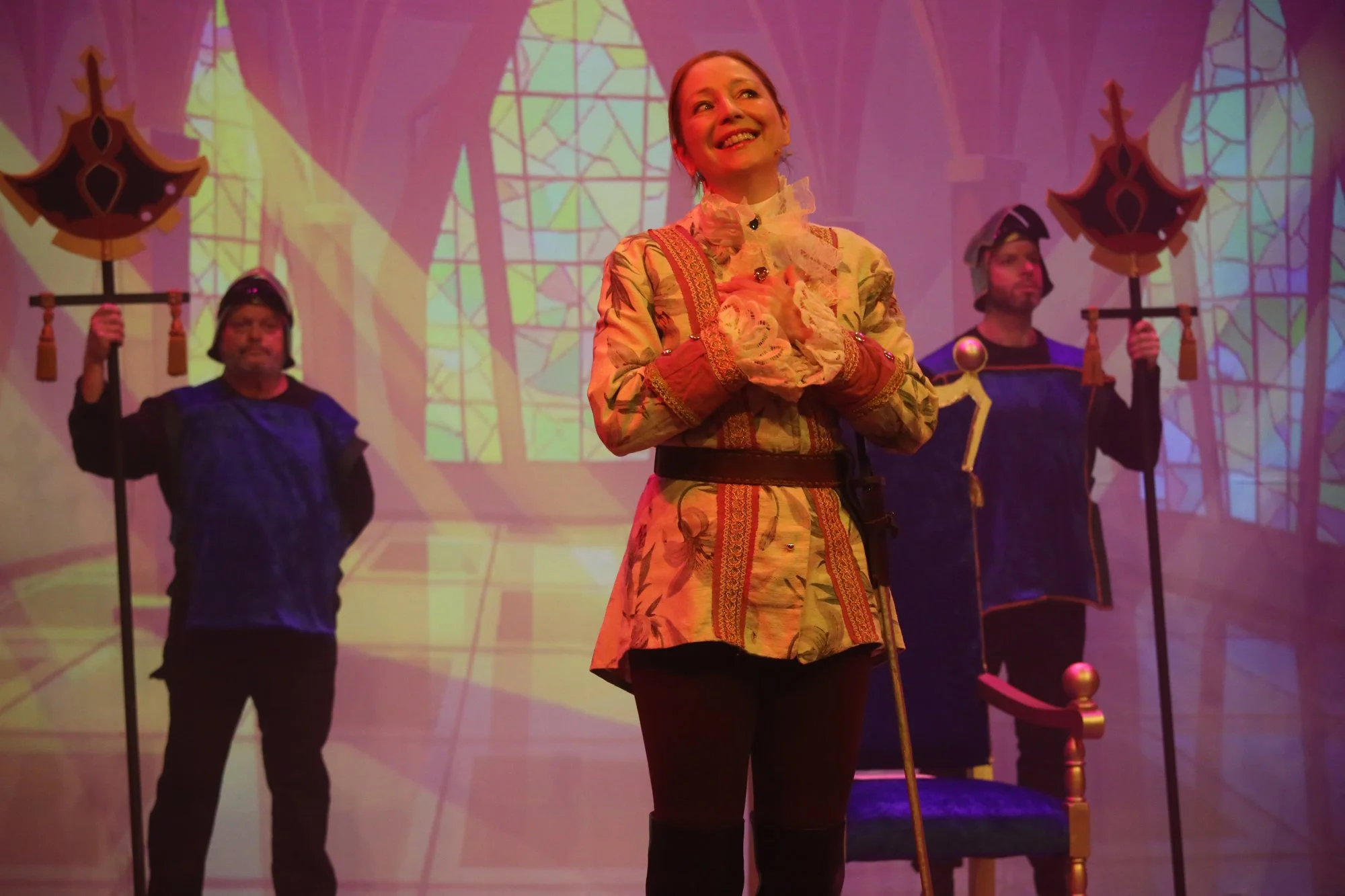 Stage scene with woman in ornate costume smiling, flanked by two men in dark clothing holding staffs with decorative tops, in front of a stained glass window backdrop.