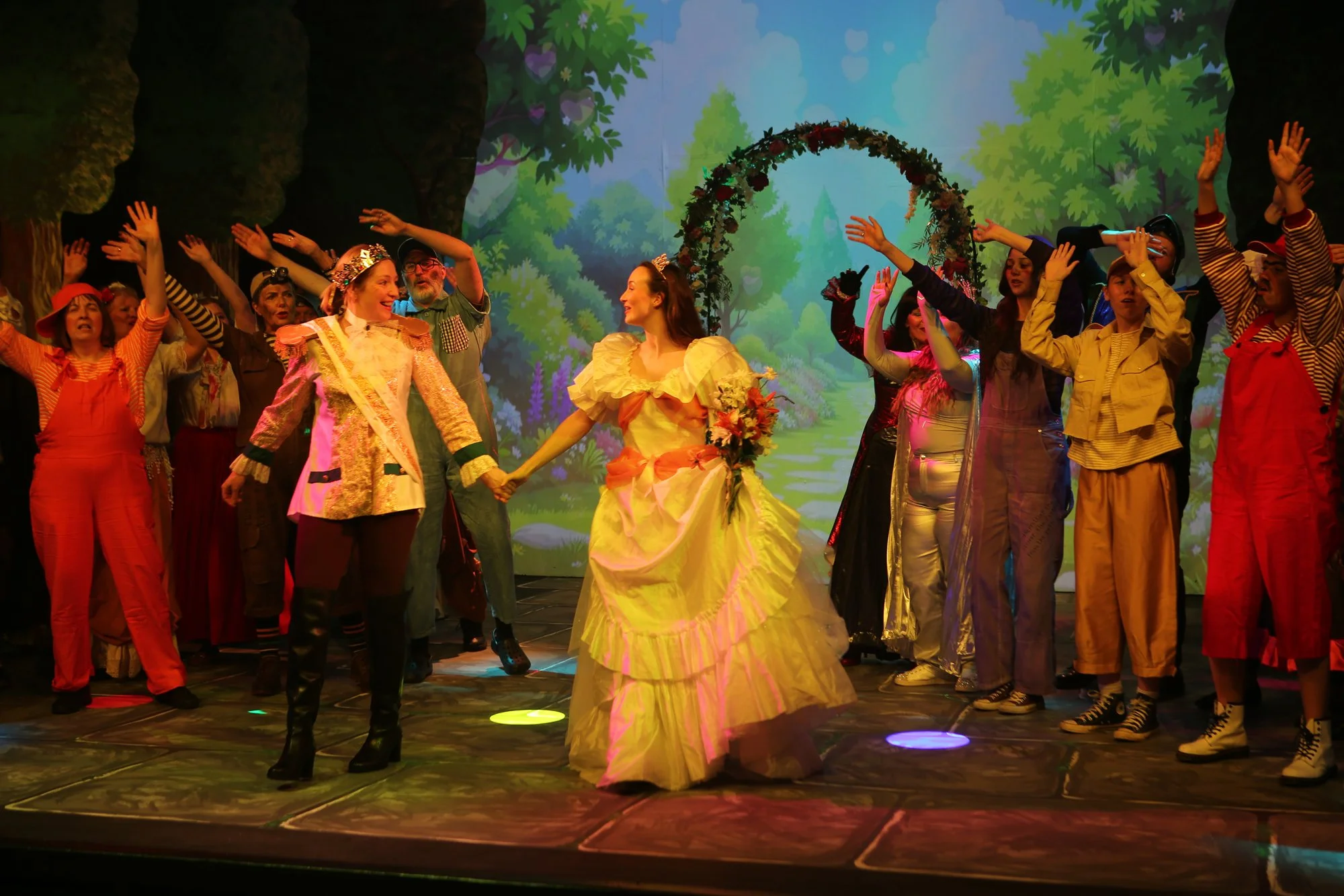 A scene from a theatrical production featuring a woman in a yellow dress holding a bouquet, standing in front of a group of performers in colorful costumes, with a backdrop of a forest scene.