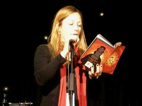 Scarlett Thomas reading a book into a microphone on a stage with dark background and small lights.
