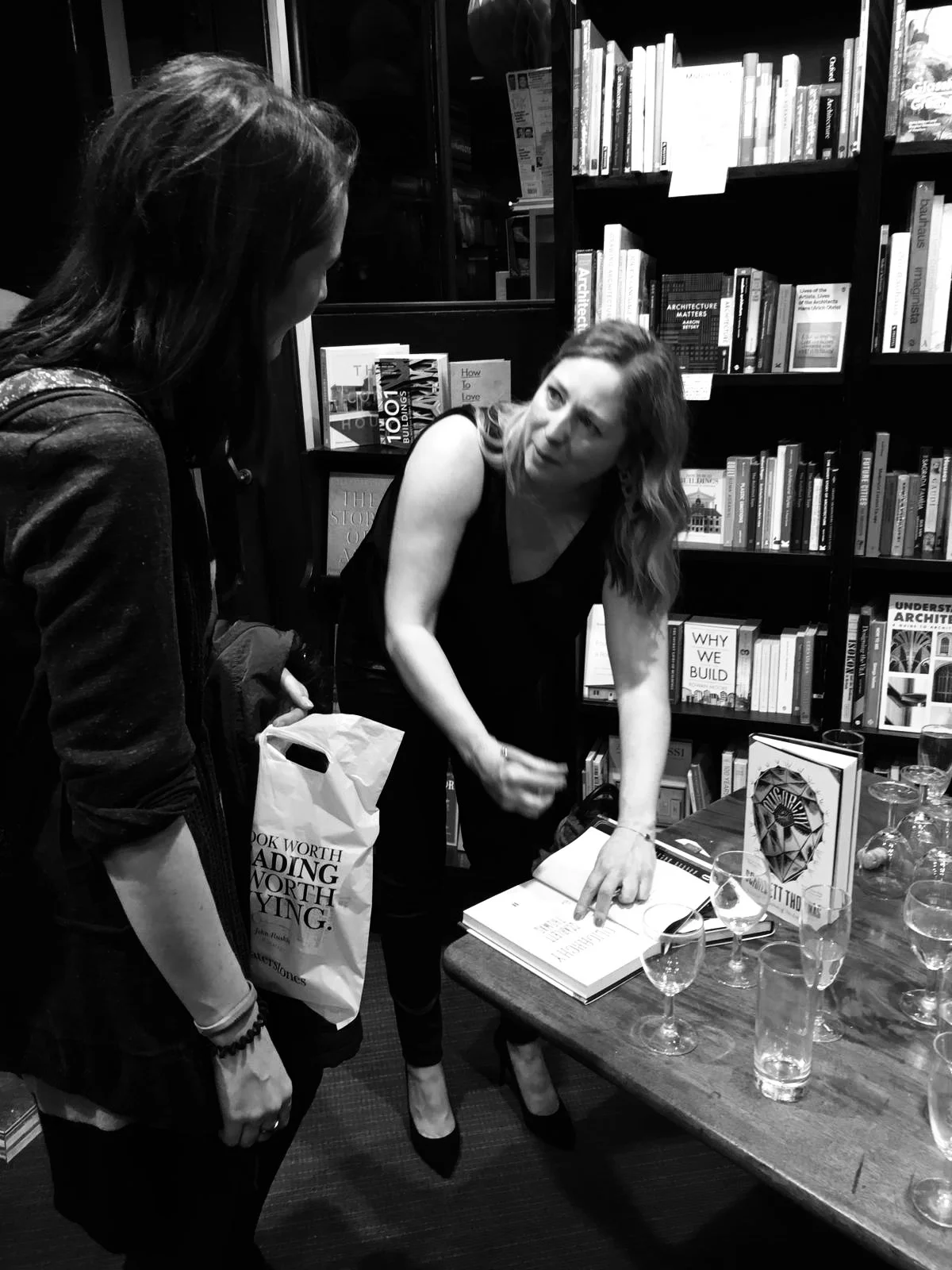A woman with shoulder-length hair, wearing a sleeveless top, points to a book on a table at a book signing event. She is signing a copy of her book while another woman, holding a tote bag with the quote 'A book worth reading is worth buying,' listens