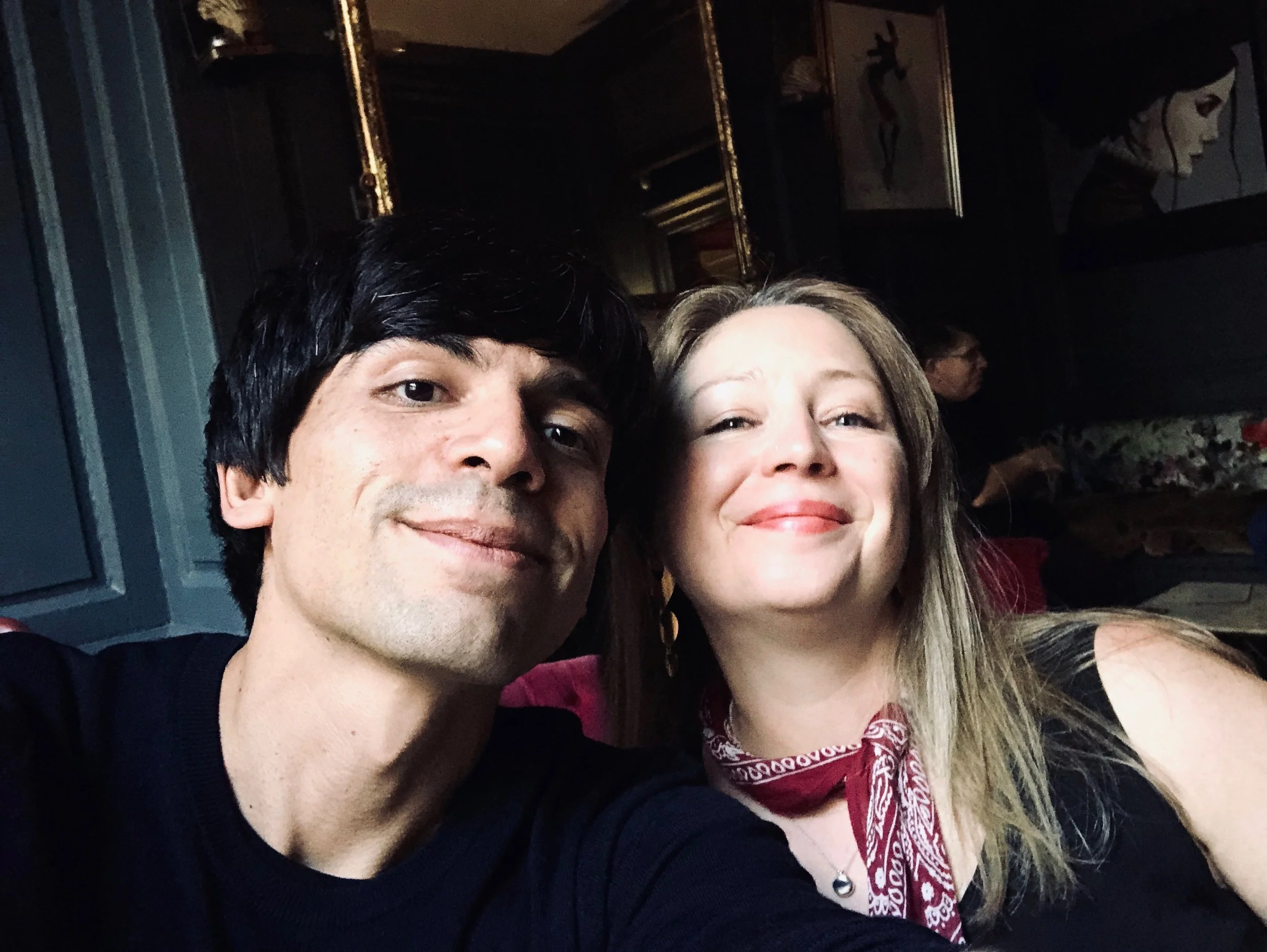 A man and woman taking a selfie together in a dimly lit room with dark decor, paintings on the wall, and a person in the background.
