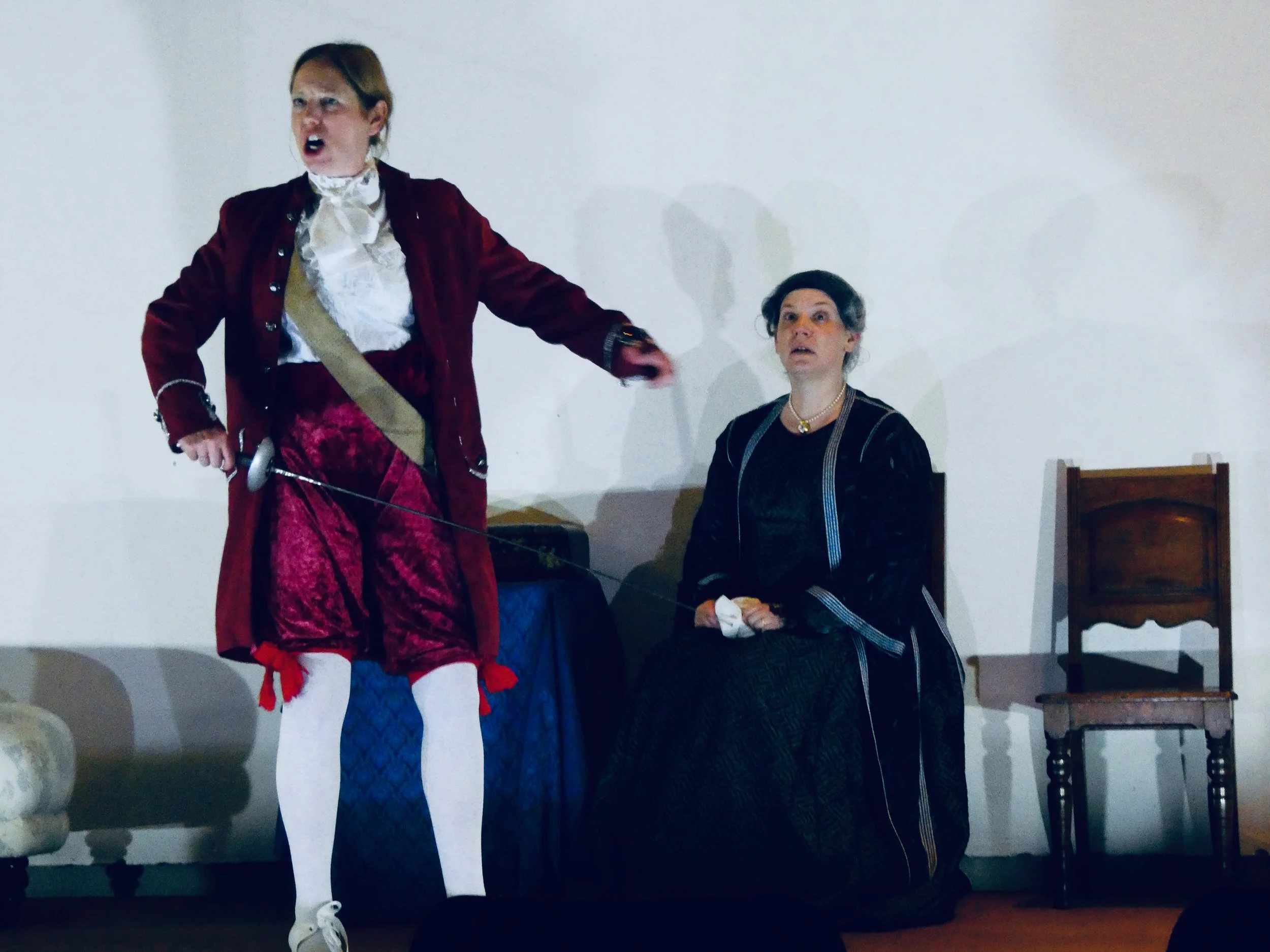 Two women dressed in historical costumes performing in a theatrical play. One woman stands, holding a sword, with an angry expression. The other woman sits on a chair, looking surprised or shocked.