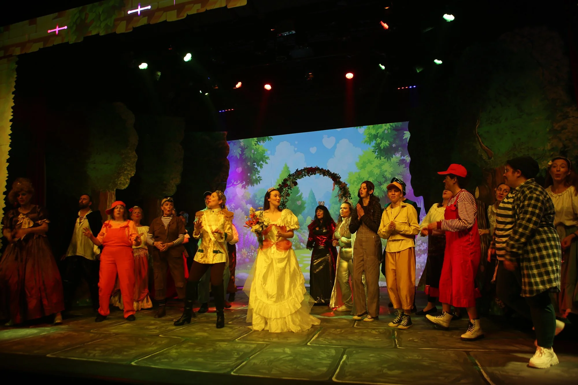 A stage scene from a theatrical performance featuring actors dressed in colorful costumes representing various characters. The background has a animated nature scene with trees and a blue sky. The actors are standing in a line, some holding hands, wi