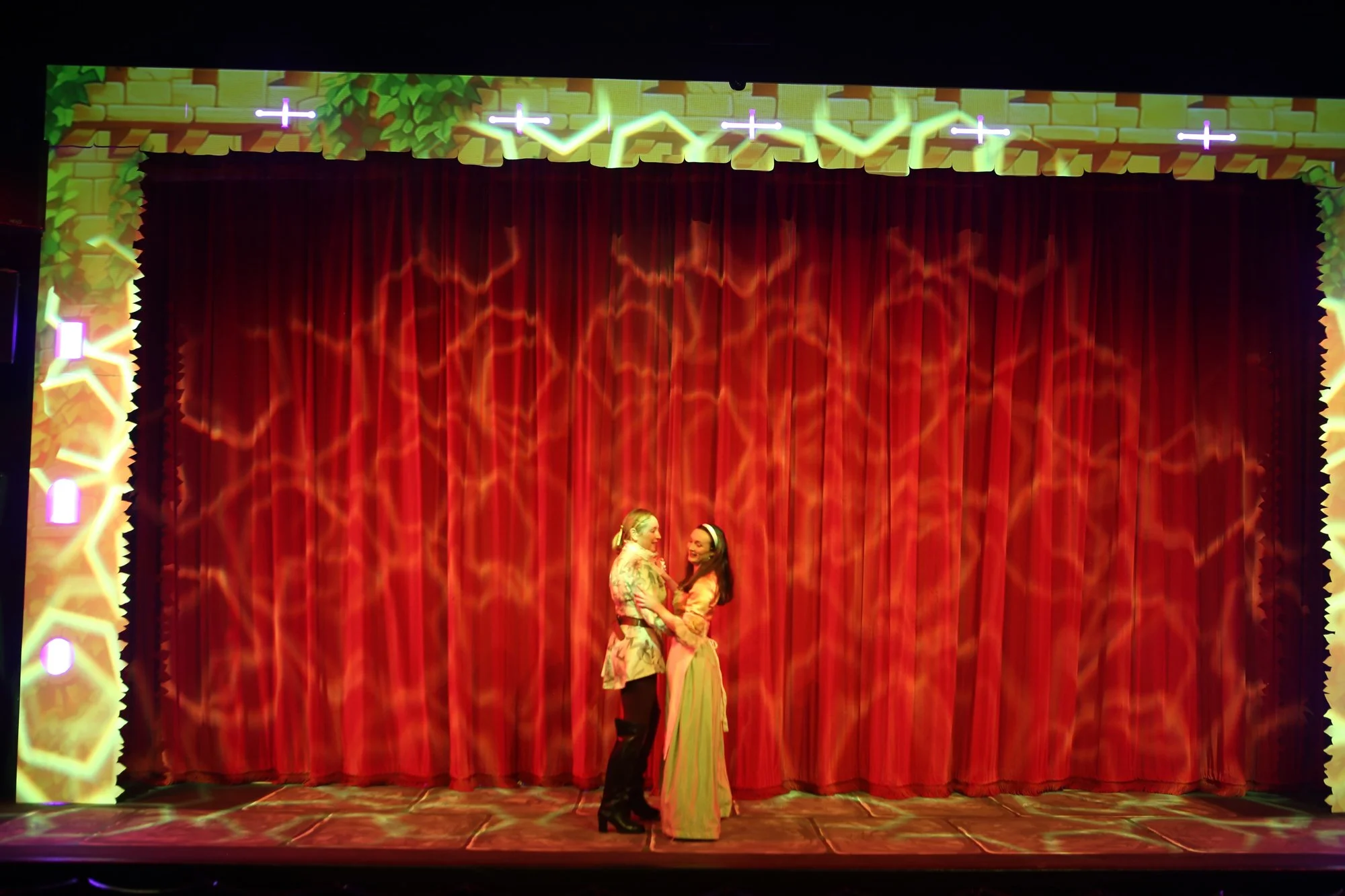 Two women standing on a red stage with a decorated backdrop, appearing to be about to kiss or embrace each other.