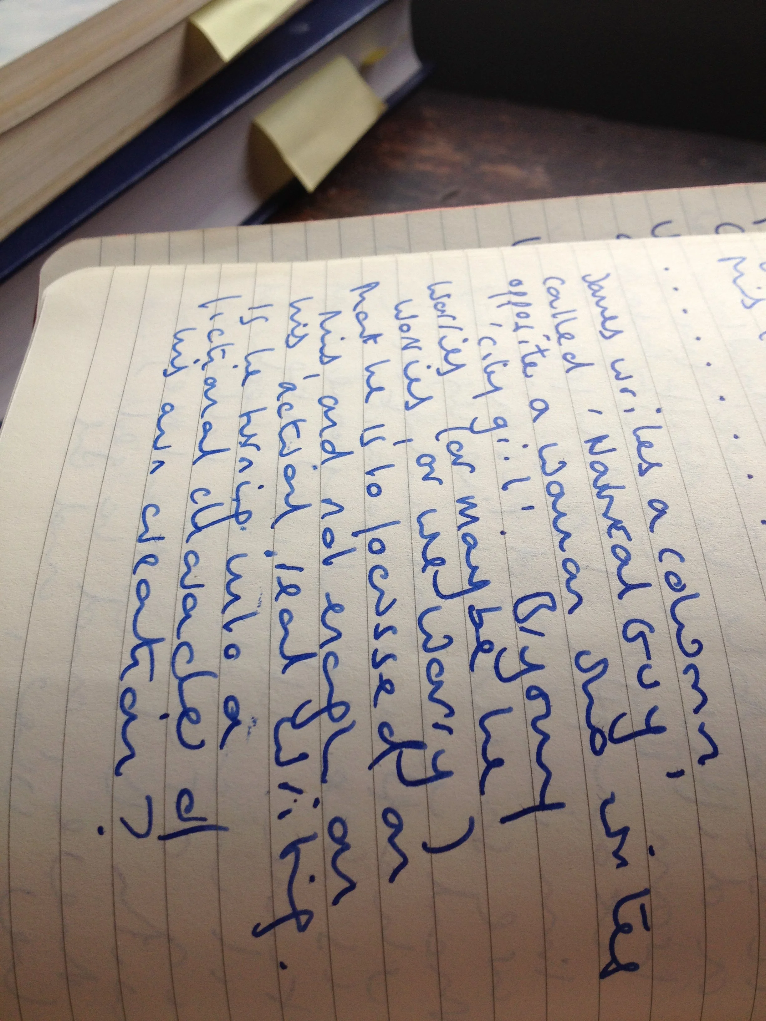 Close-up of a handwritten note in blue ink on lined paper, resting on a desk with a book and sticky notes in the background.