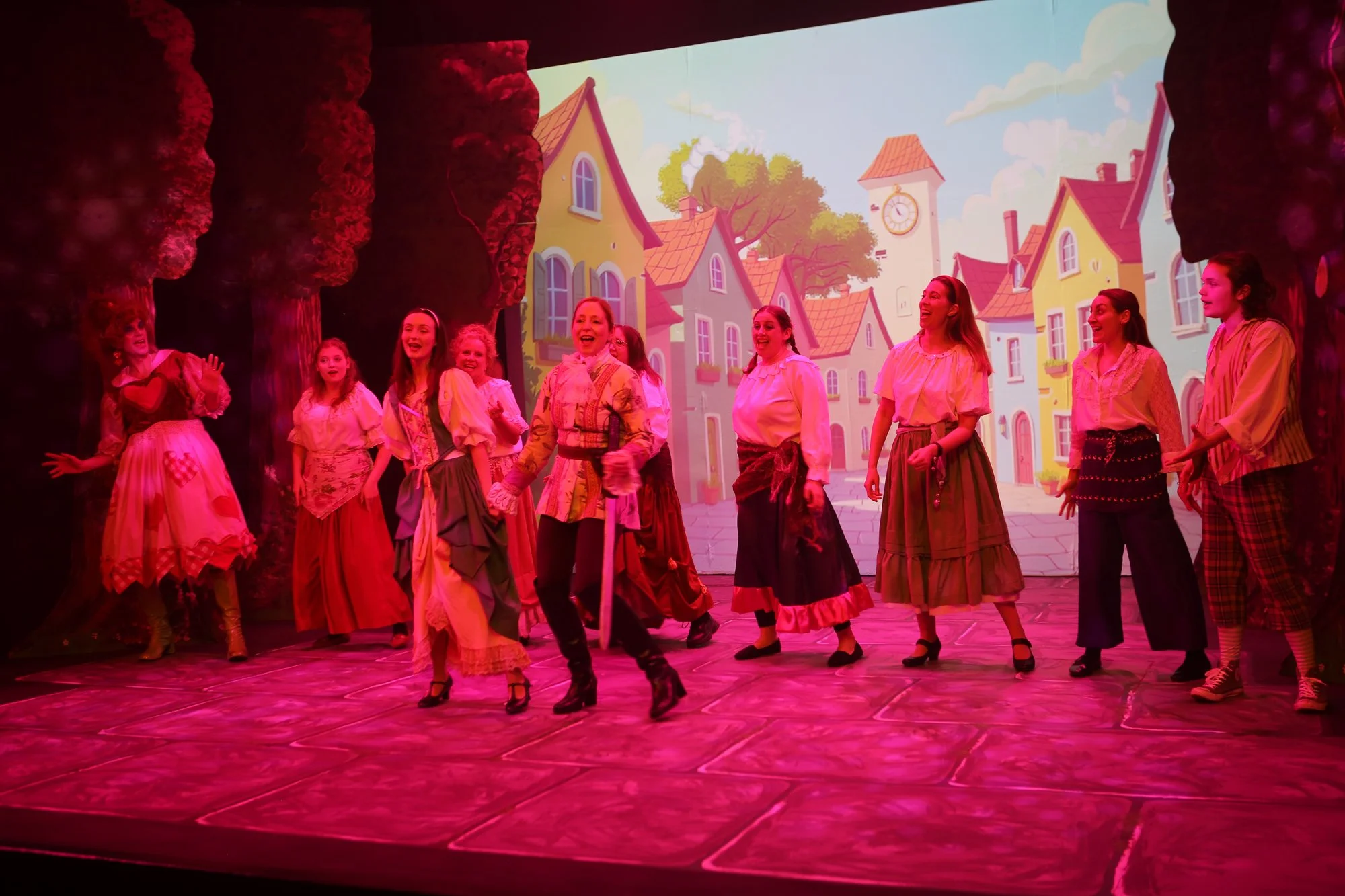A group of women and girls performing a stage play in costumes, with a colorful animated village scene projected in the background.