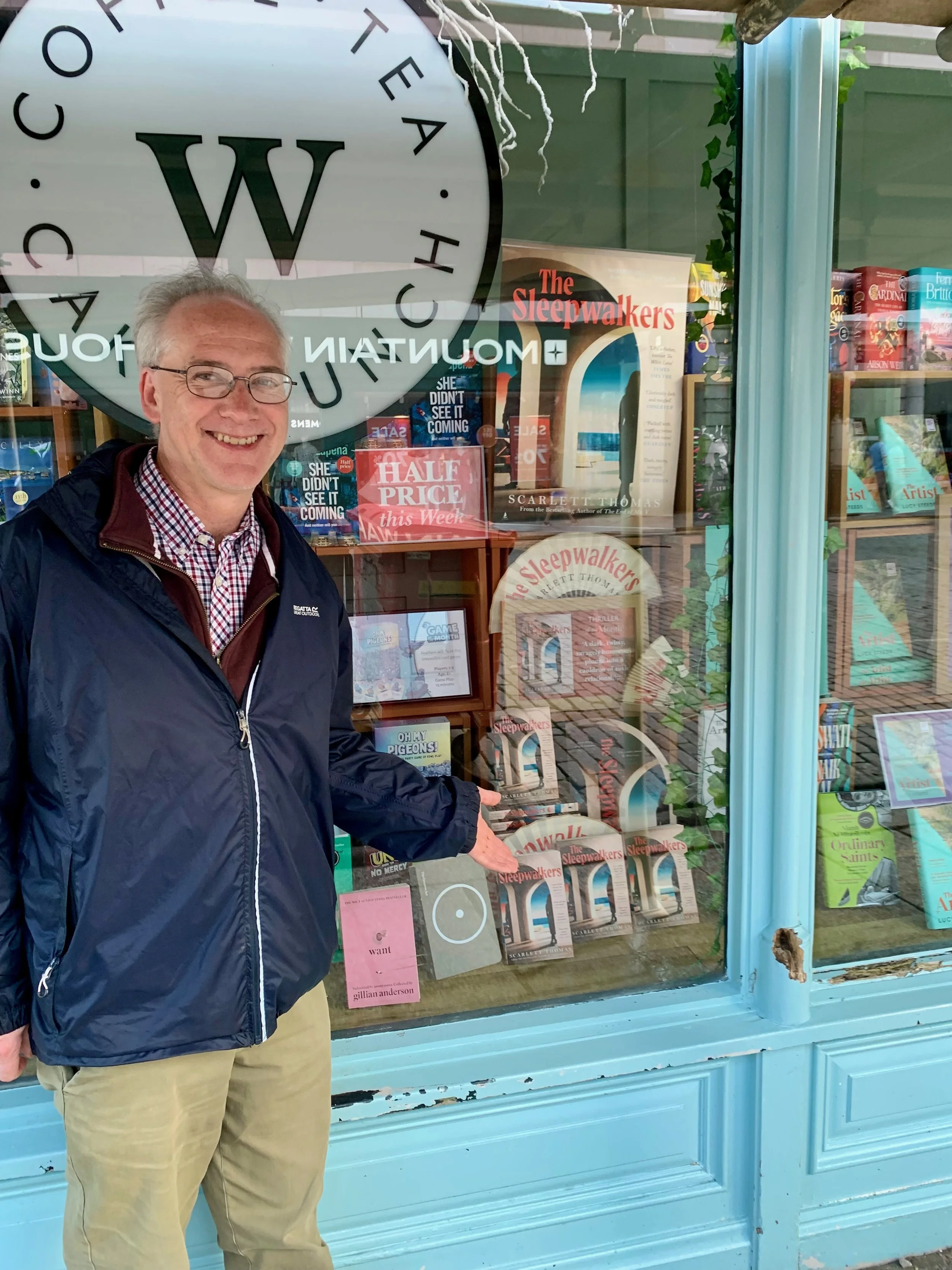 A man standing outside a bookstore window, smiling and pointing at books inside, with the store's logo on the window.