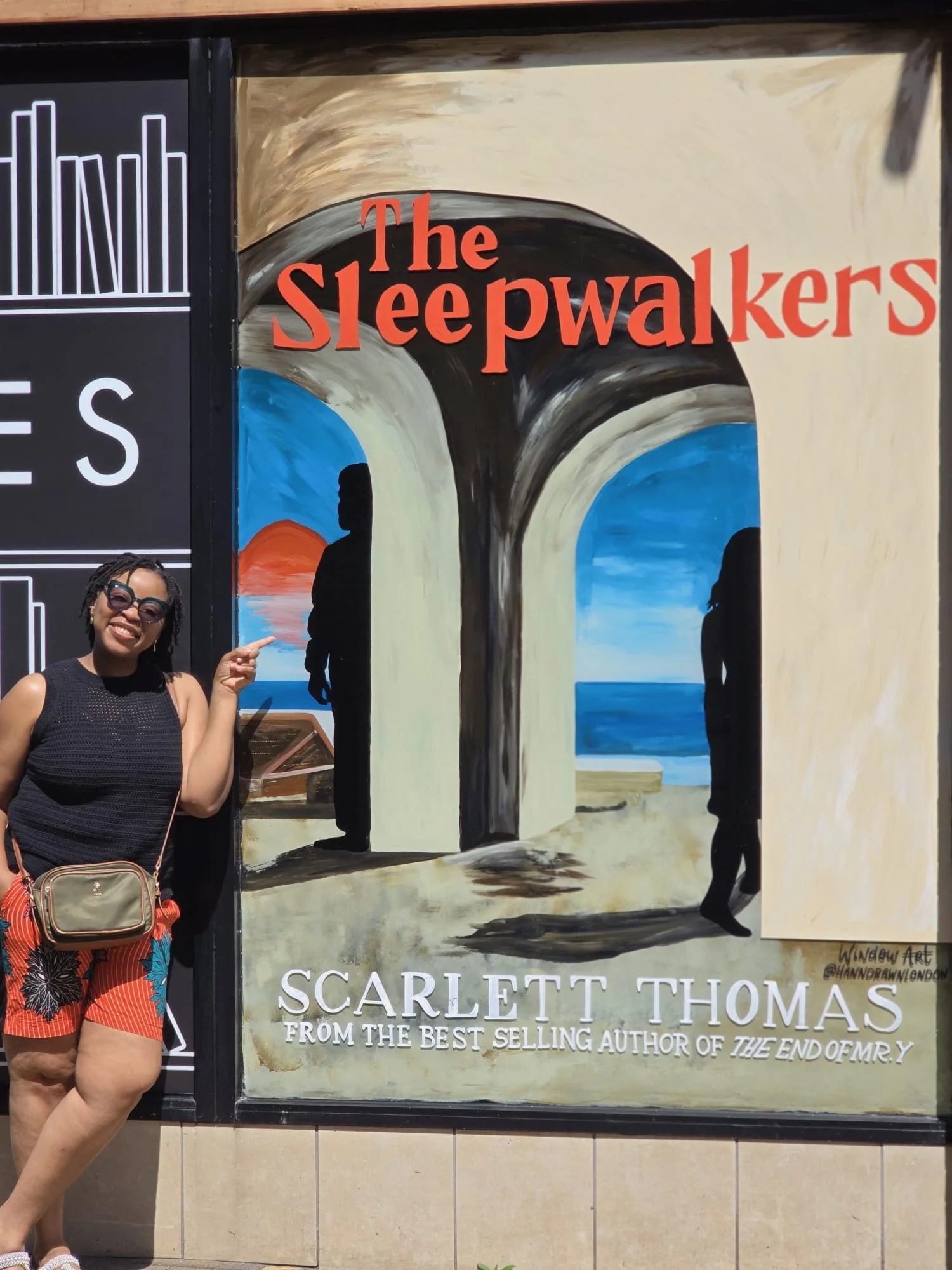A woman standing next to a mural advertising the book 'The Sleepwalkers' by Scarlett Thomas. The mural depicts a harbor scene viewed through a stone arch, with a silhouette of a person and a sunset over the sea.