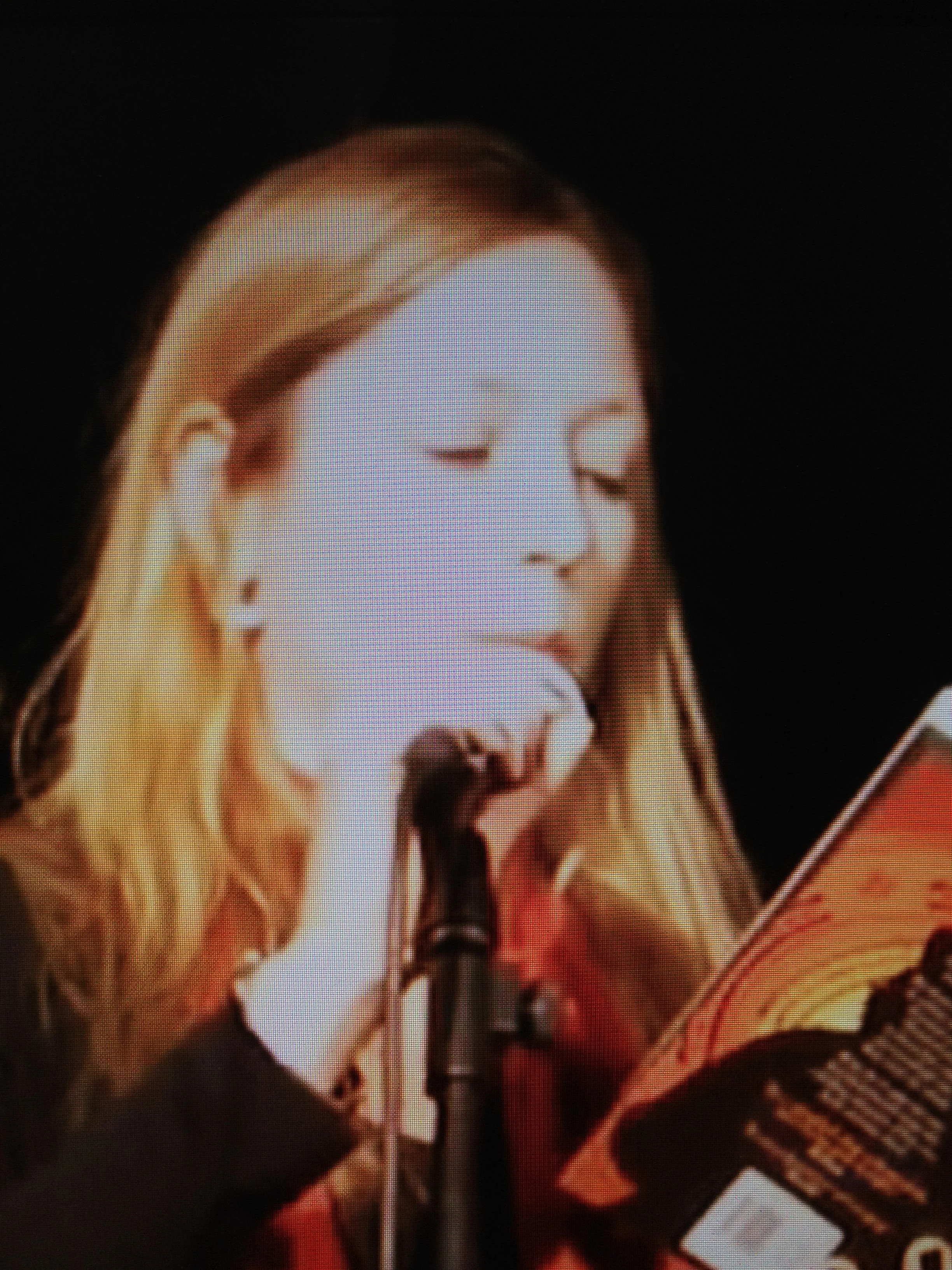 A young woman with blonde hair and earrings is reading a book or magazine in front of a microphone with her hand resting on her chin.
