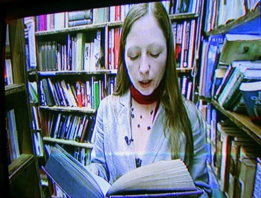 A woman with long hair and a red choker reading a book in a library with bookshelves in the background.