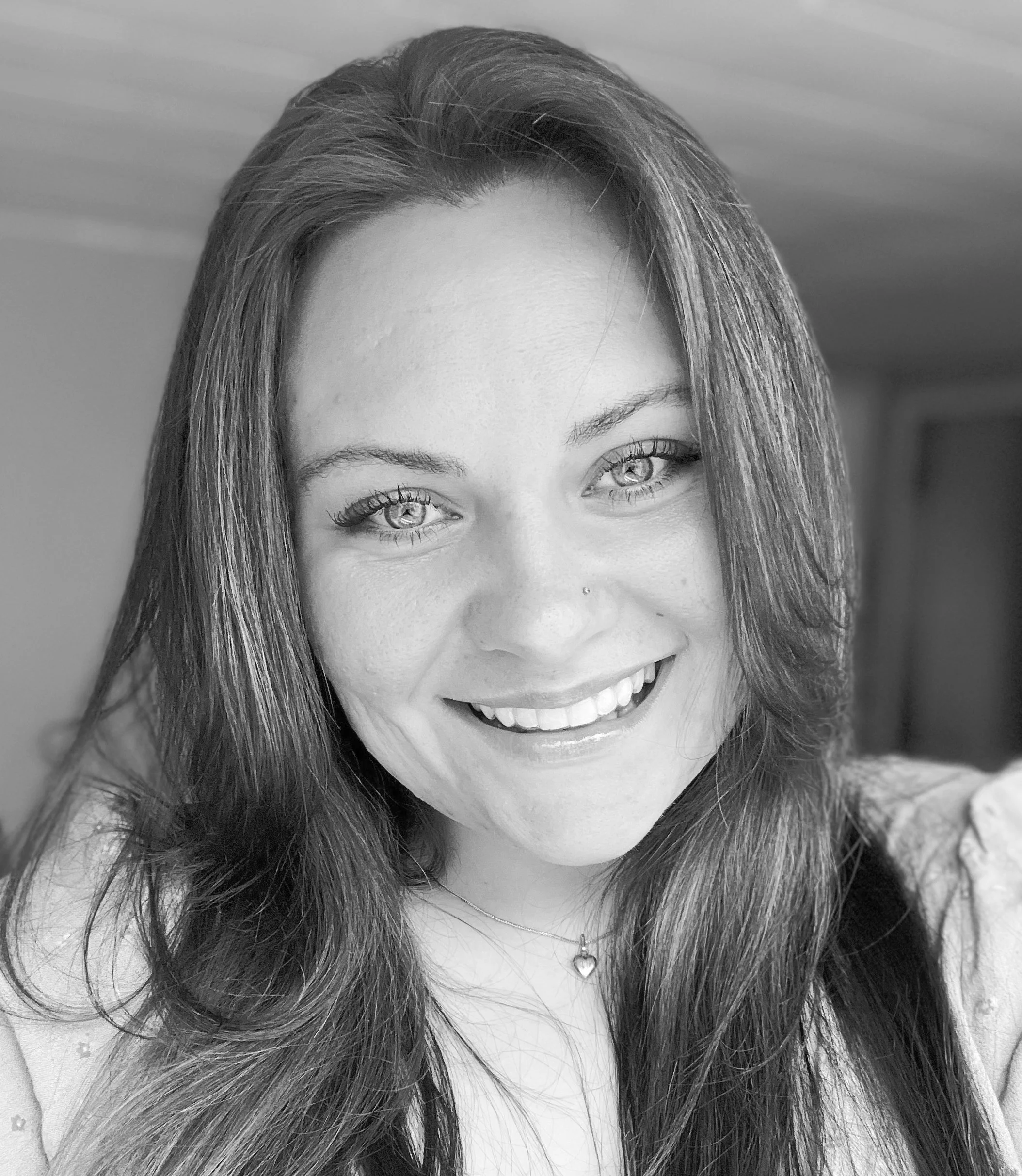 A smiling woman with long dark hair, light eyes, and a nose piercing, wearing a necklace with a heart pendant.