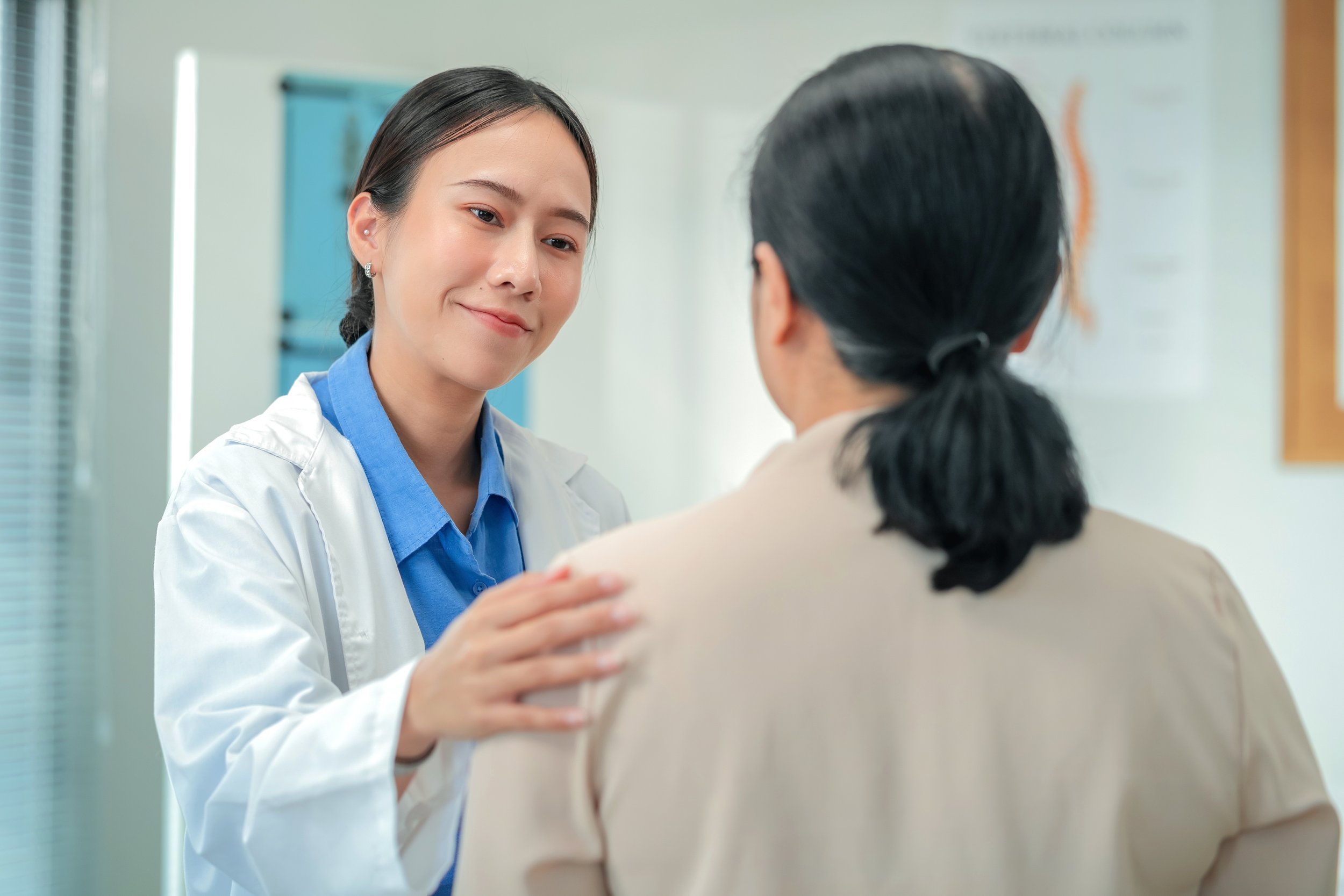 A young female doctor with dark hair and earrings smiling and comforting a patient in a hospital or clinic setting.
